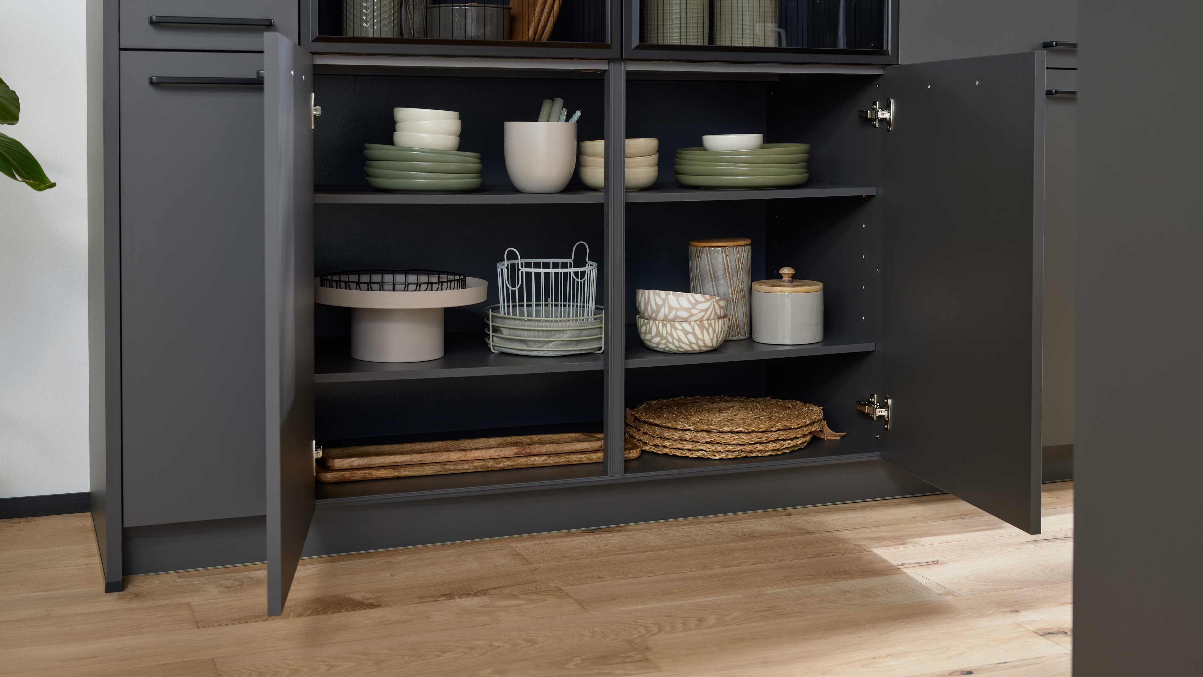 Dunkelgraues offenes Küchenschrank-Unterteil mit zwei Türen und Regalen, gefüllt mit Tellern, Schalen, Dosen, Korb und Holzbrett.