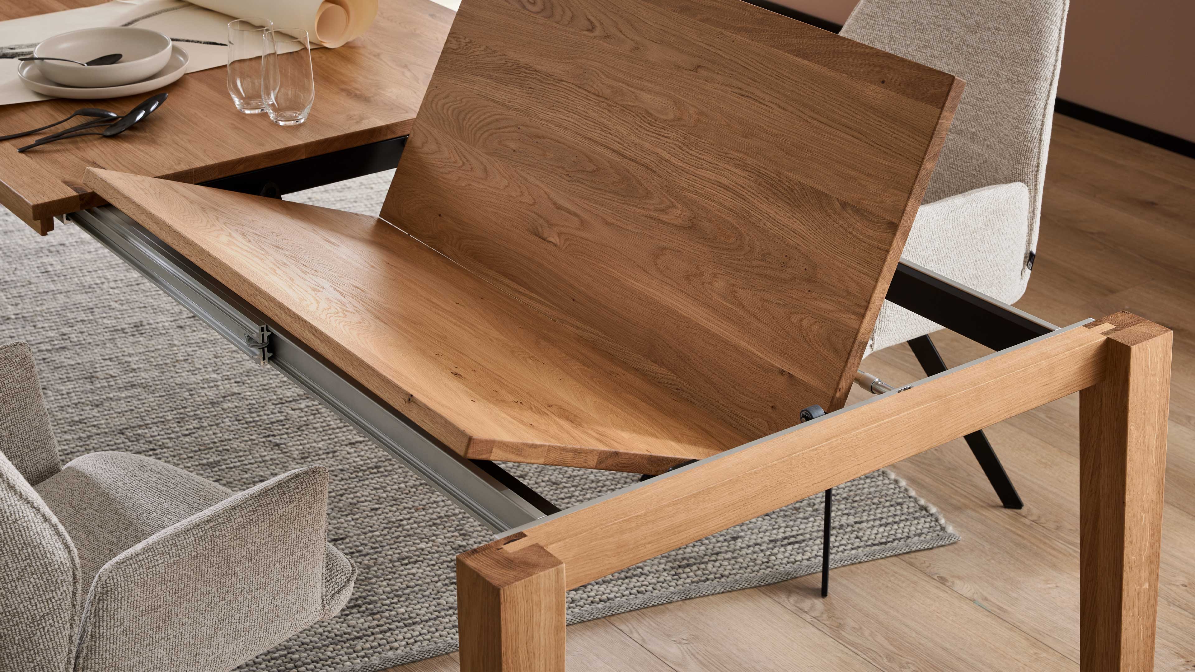 Ausziehbarer Esstisch aus hellem Holz mit aufgeklappter Einlegeplatte auf Metall-Schienen, zwei gepolsterte Stühle sichtbar.