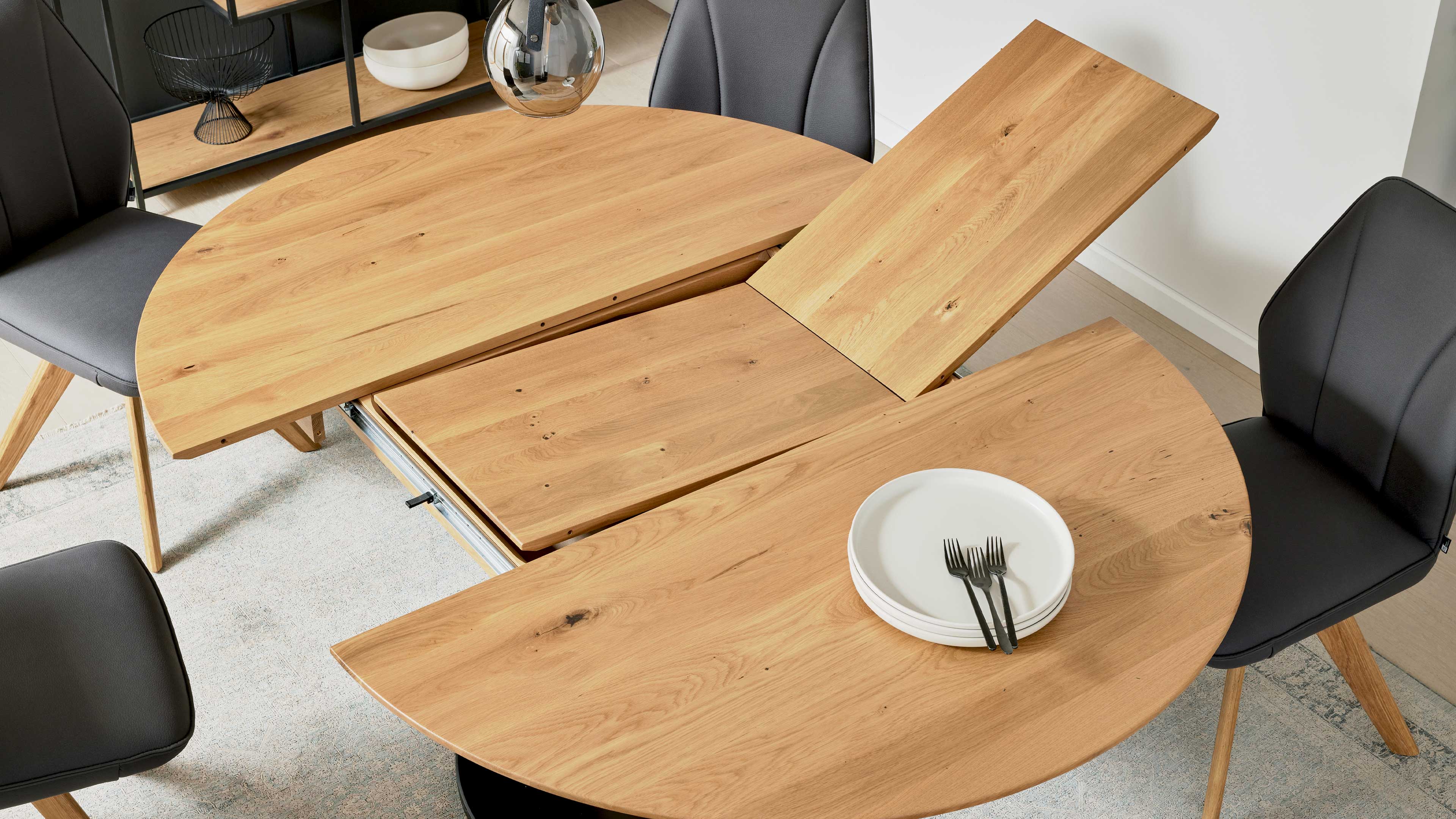 Runder, ausgezogener Holztisch mit Verlängerungsplatte, darauf weiße Teller und Gabeln, umgeben von dunklen Stühlen.