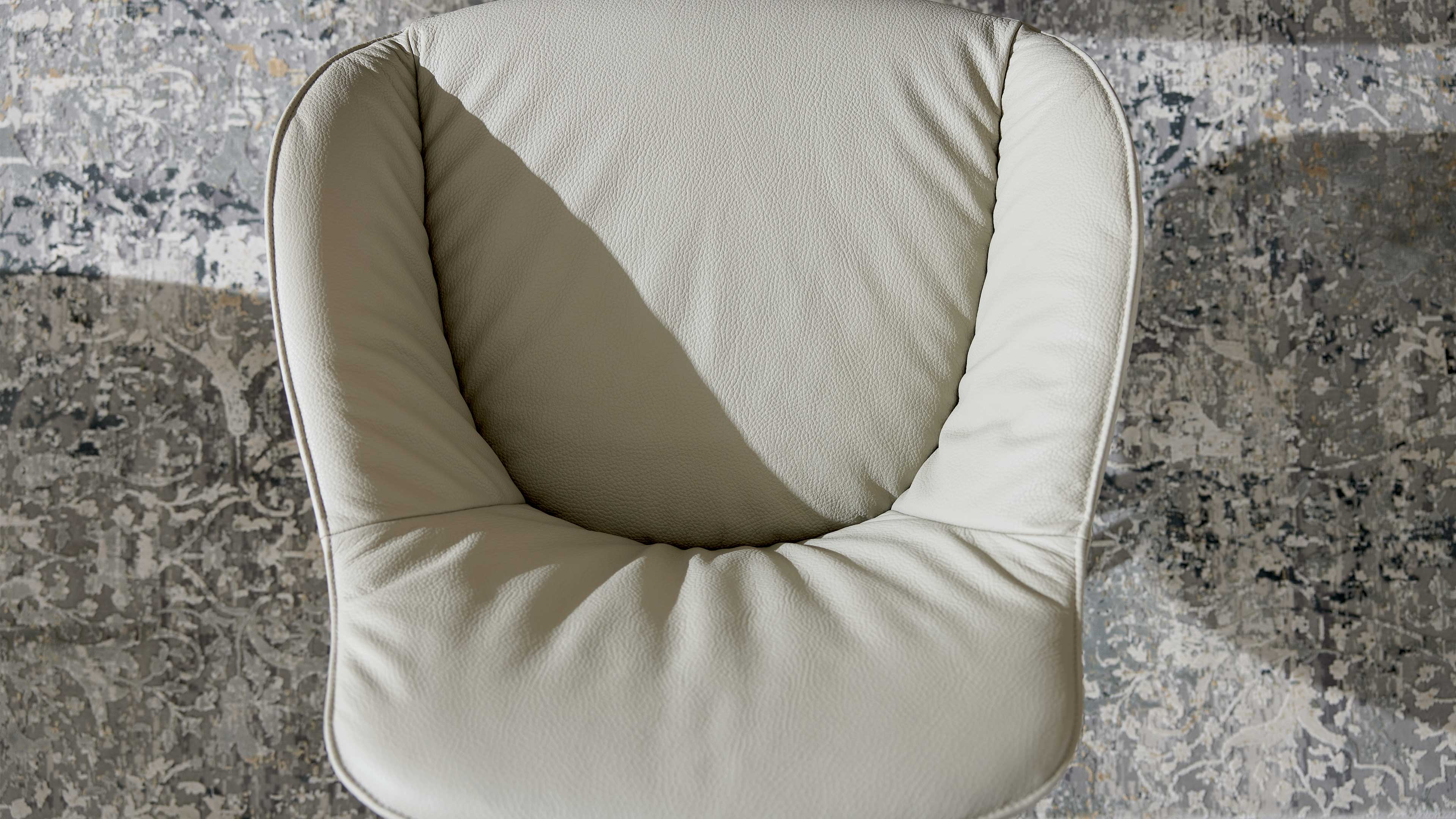 Beige gepolsterter Ledersessel von oben, mit sichtbarer Narbung, gebogenen Armlehnen, Dreiecks-Schatten und gemustertem Teppich darunter.