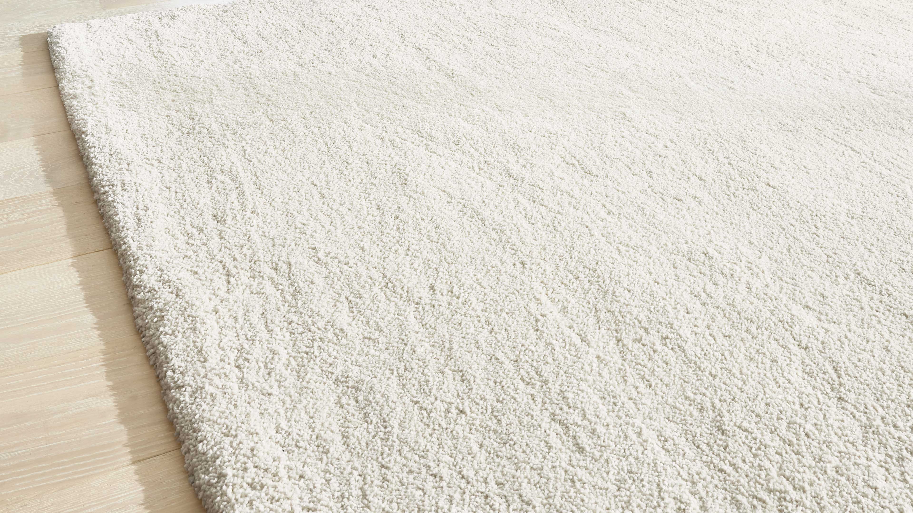 Cremeweißer Hochflor-Teppich mit dichter Textur bedeckt fast die gesamte Fläche, links ist die Kante auf hellem Holzboden sichtbar.
