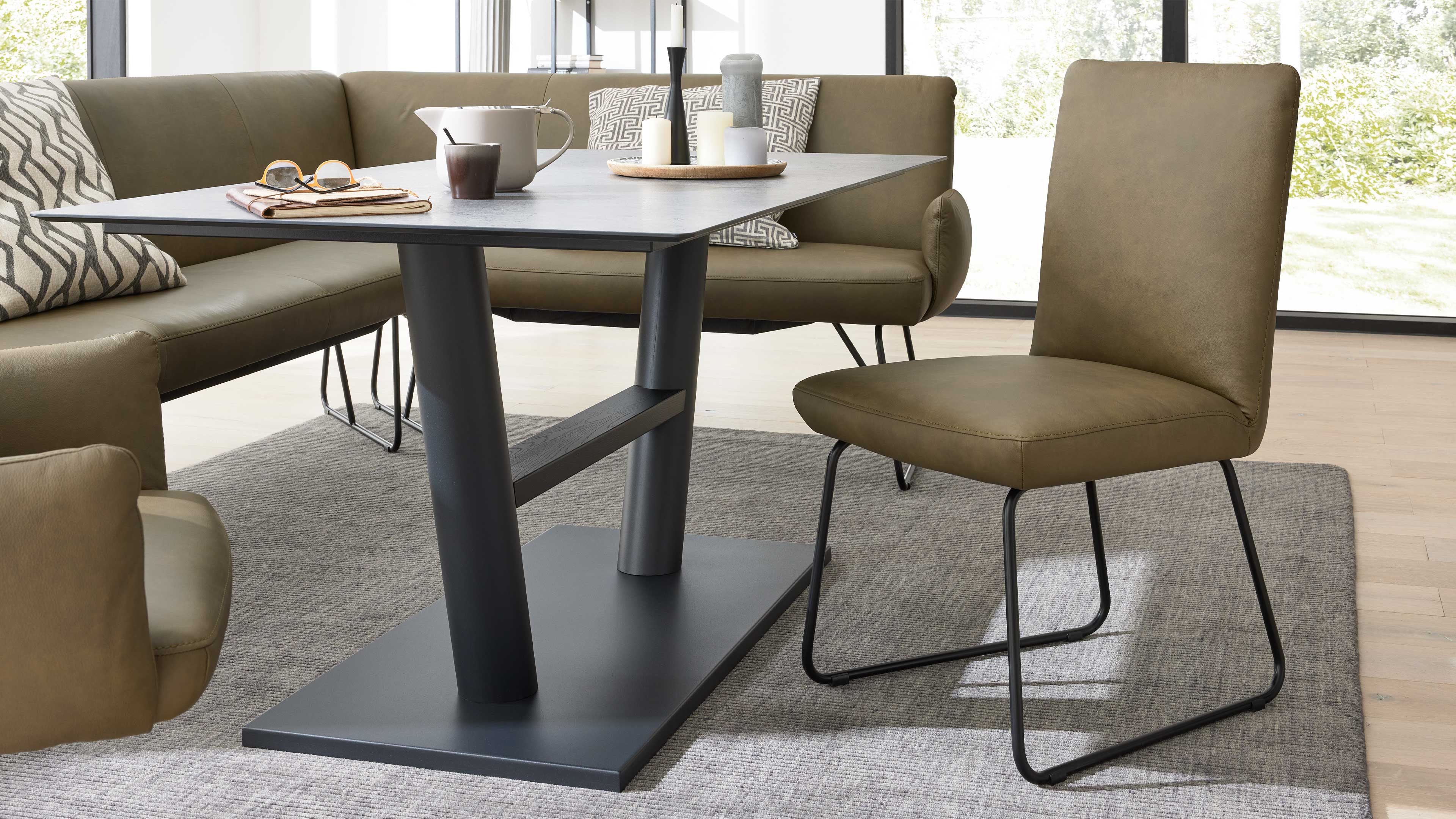 Olivefarbener Stuhl mit schwarzer Kufen‑Metallkonstruktion neben schwarzem Tisch mit zentralem Fuß, Eckbank und Tischdeko.