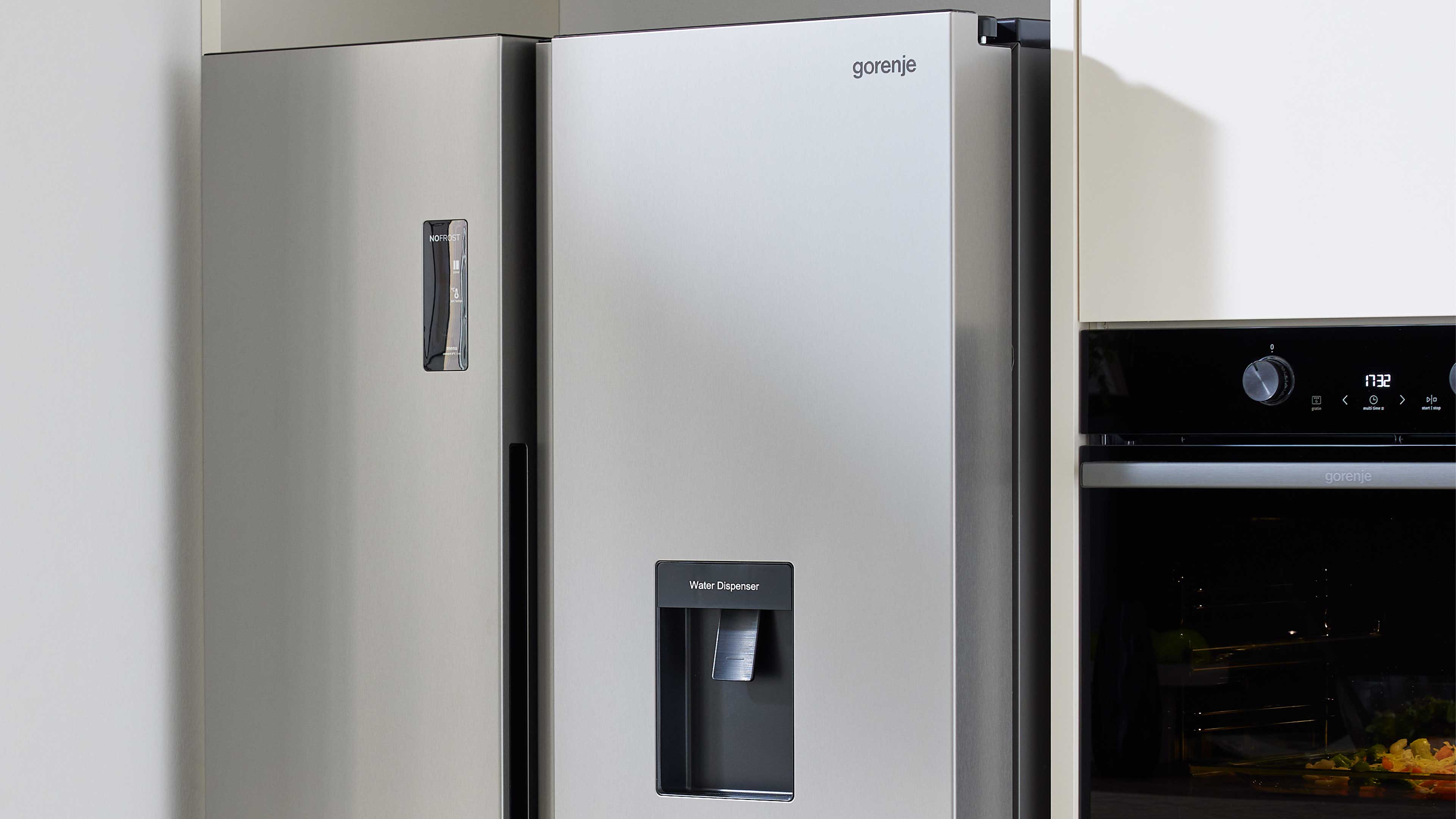 Links stehender silberner Side-by-Side-Kühlschrank mit integriertem Wasserspender und schwarzen Griffen, rechts ein eingebauter Ofen.