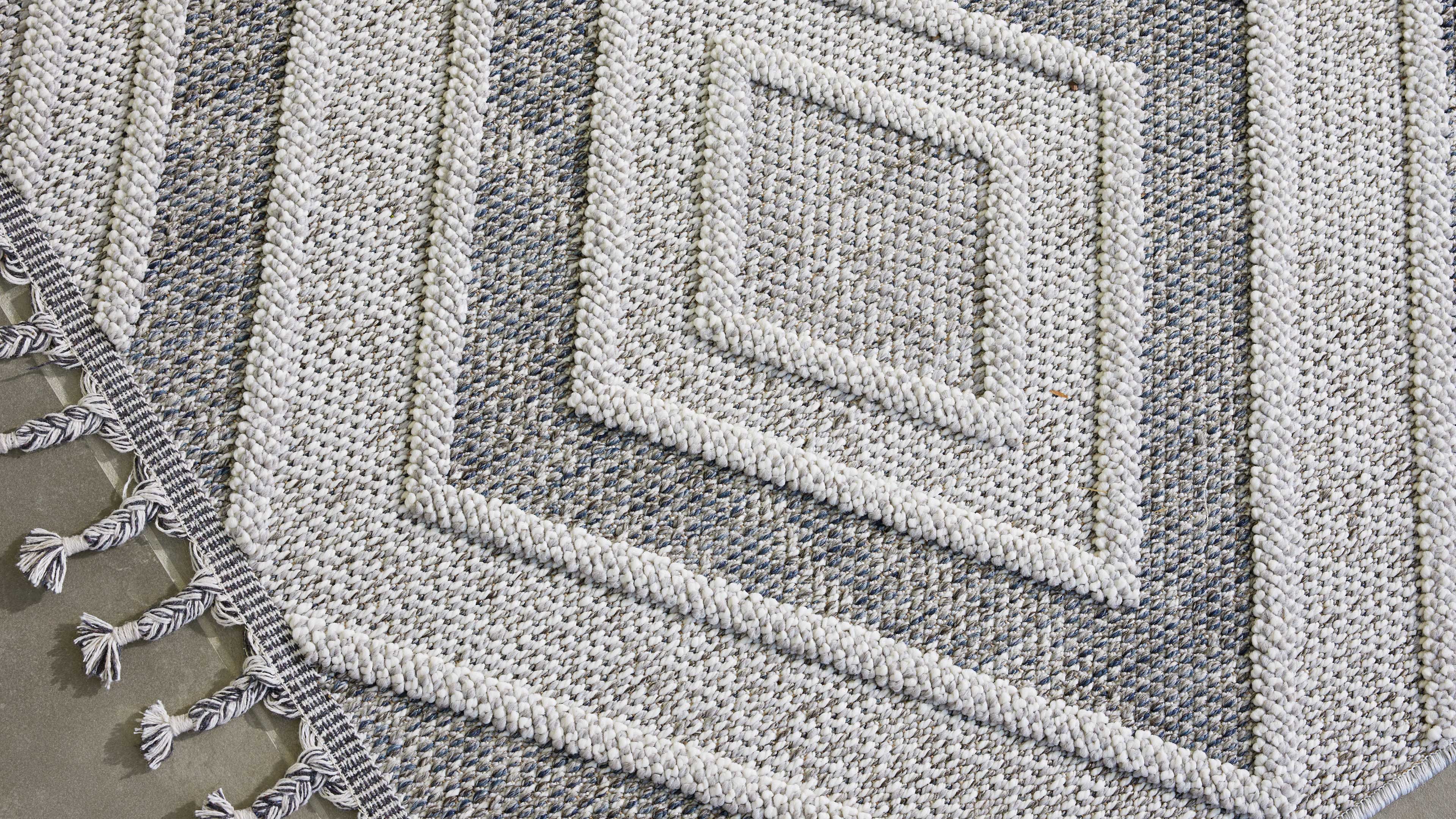 Runder Teppich mit aufgesetzten, dicken Zopfstreifen in Quadratmustern, hellgrau-blau meliertem Gewebe und Fransen an einer Kante.