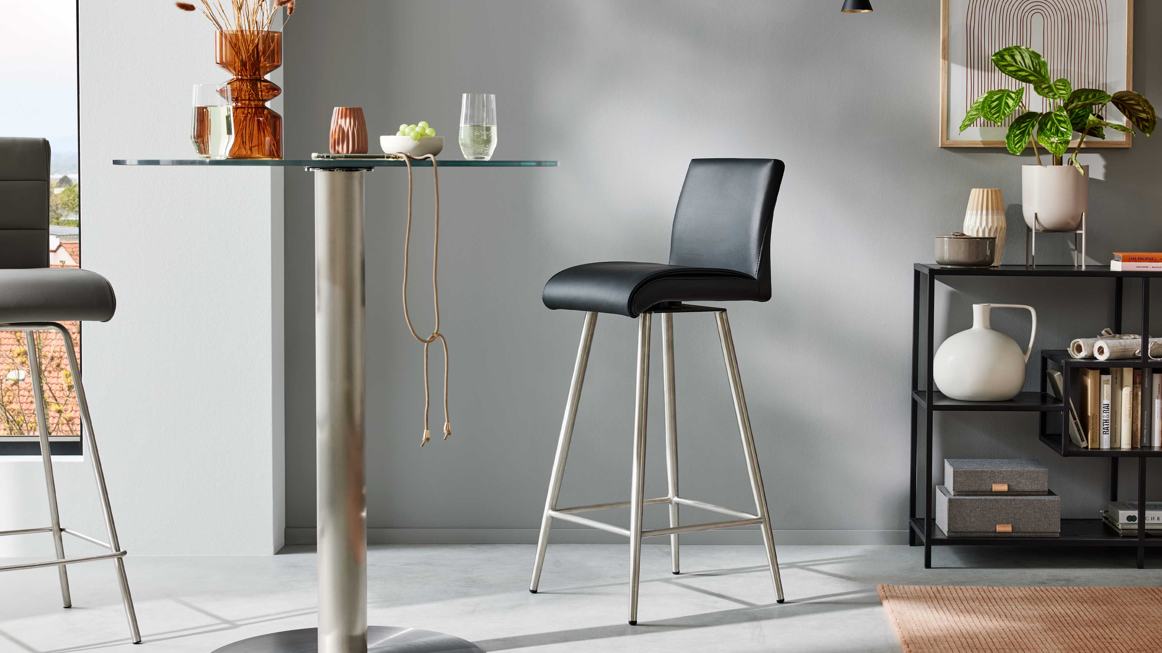 Schwarzer Barhocker mit gepolstertem Sitz auf 4 Metallbeinen vor Glas-Bartisch, links Vasen und Glas, rechts Regal mit Pflanze und Keramik.