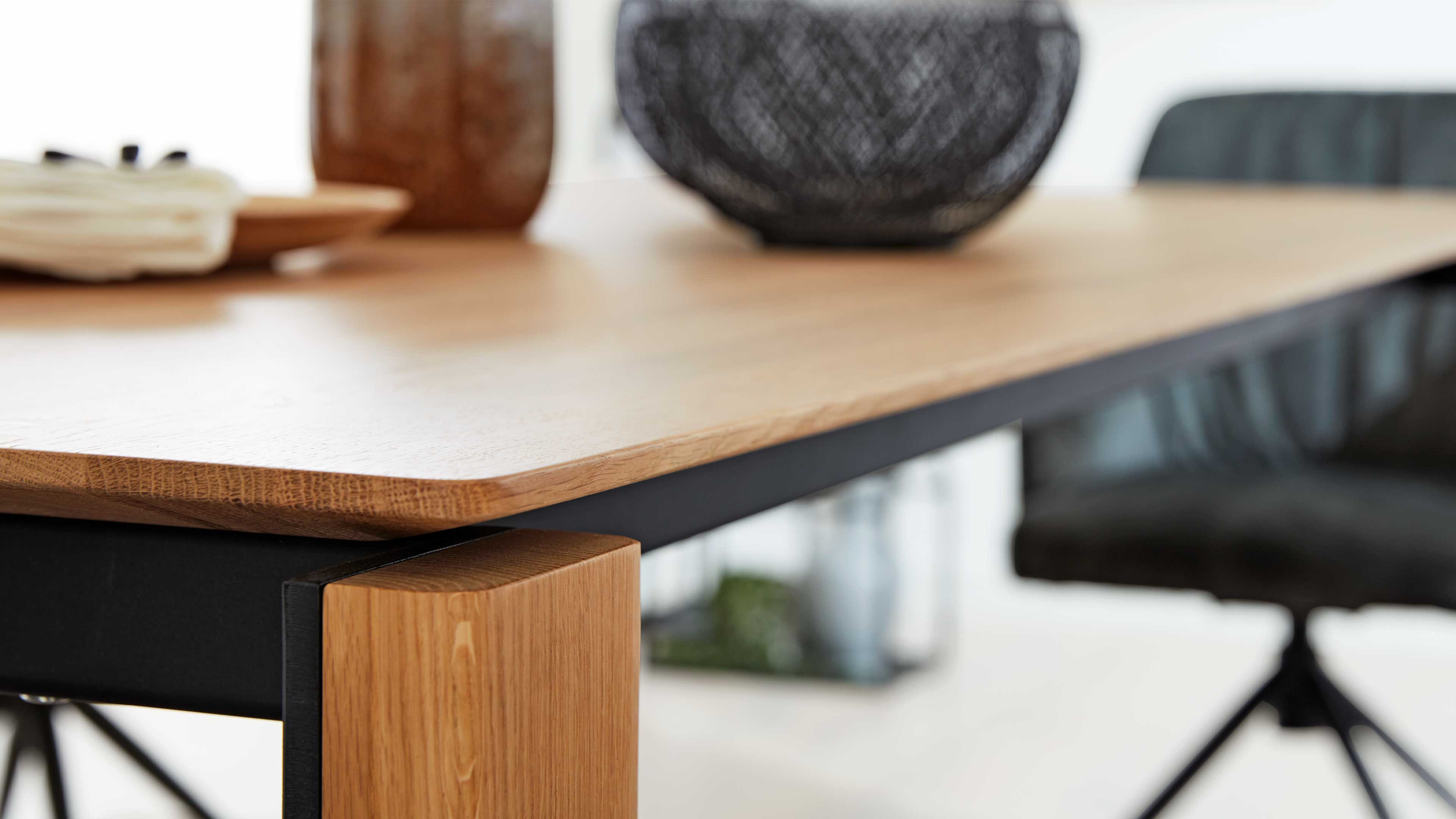 Helle Holz-Tischplatte mit schwarzem Metall-Untergestell und eckigem Holzfuß, im Hintergrund Stuhl und Dekor.