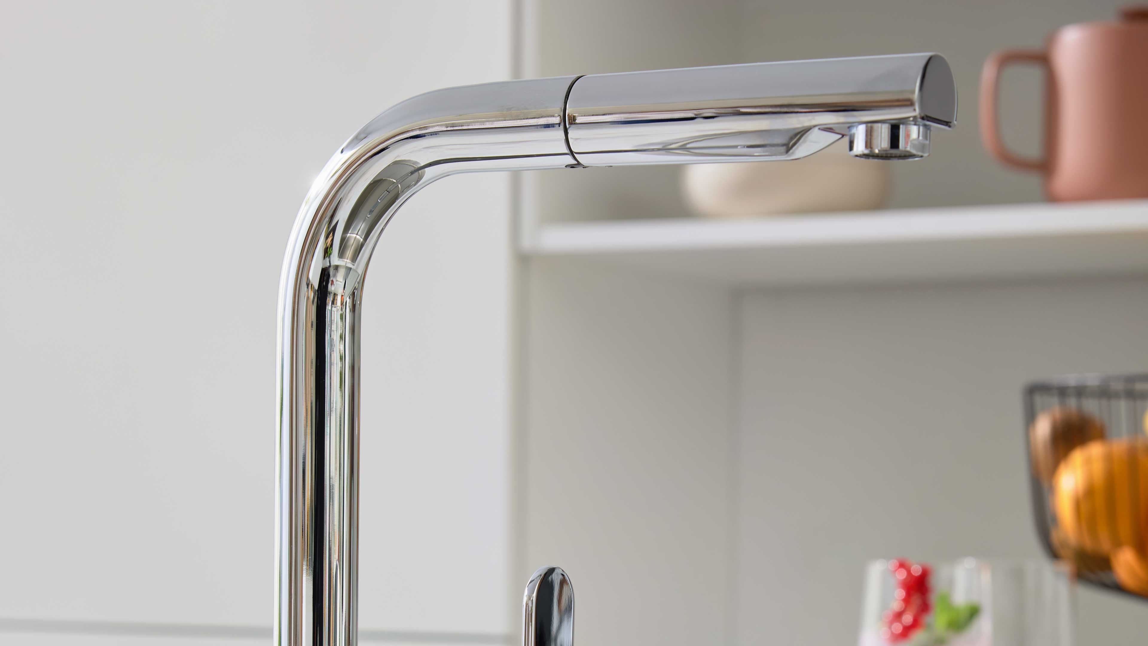 Chromfarbene Küchenarmatur mit hohem gebogenem Auslauf und Einhebelgriff, im Hintergrund unscharfe Tasse und Obstkorb.