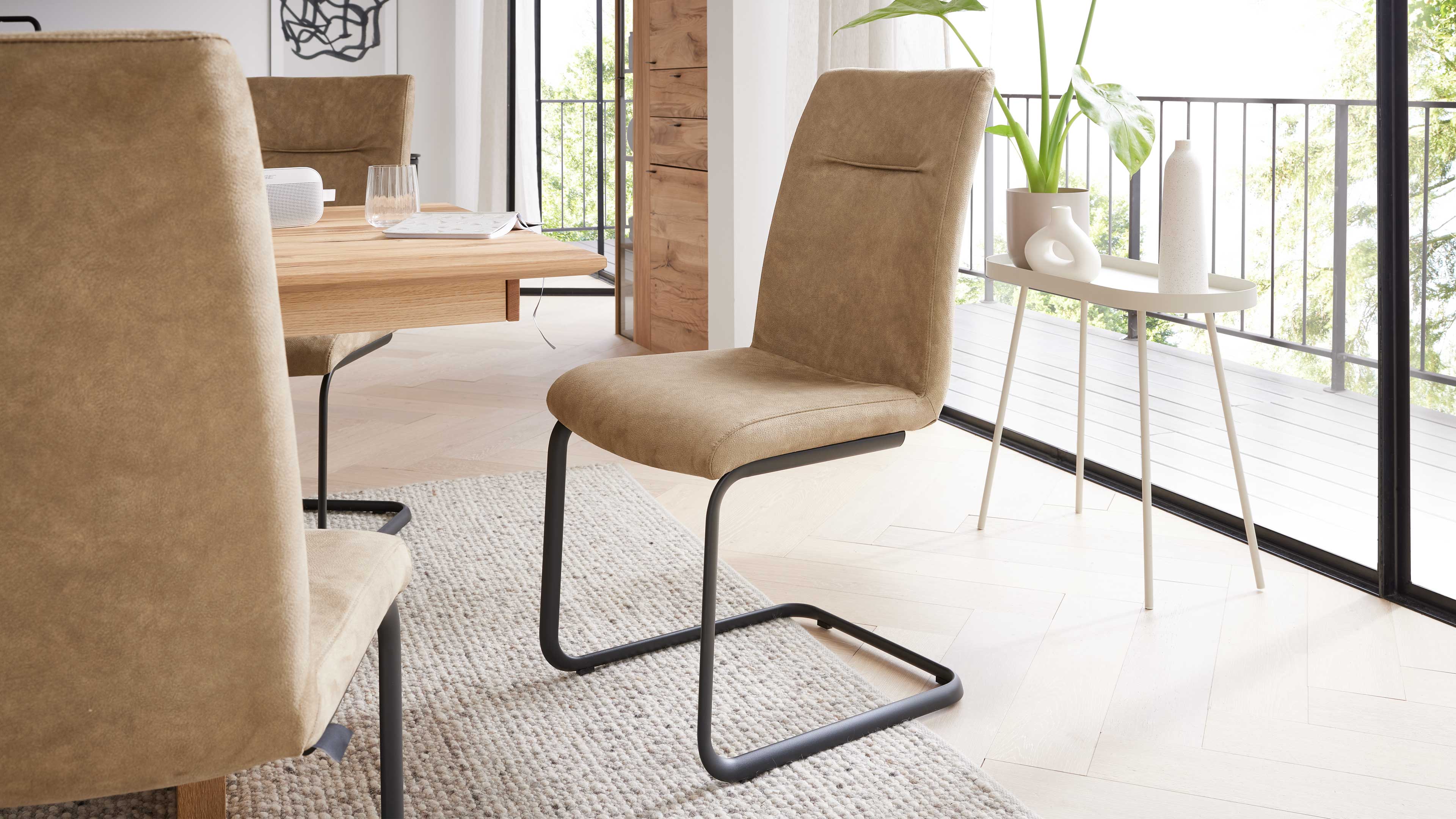 Brauner, gepolsterter Freischwinger mit schwarzem Metallgestell an einem Holztisch, daneben Beistelltisch mit Pflanzen und Vasen.