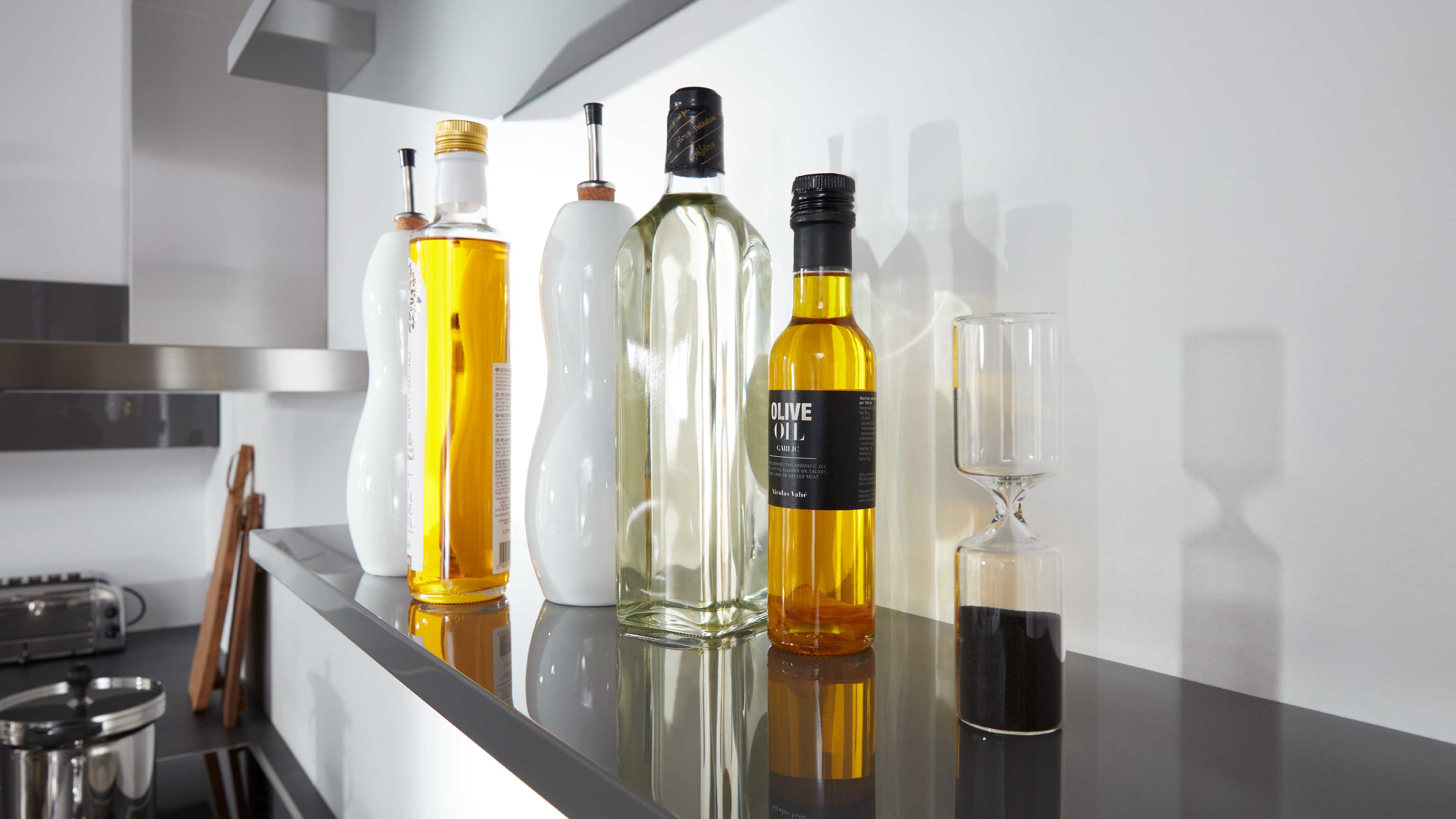 Schlanke Glasflaschen mit gelb-goldenem Öl, zwei weiße geschwungene Keramikspender und eine Glasuhr mit schwarzem Sand auf schwarzem Regal.