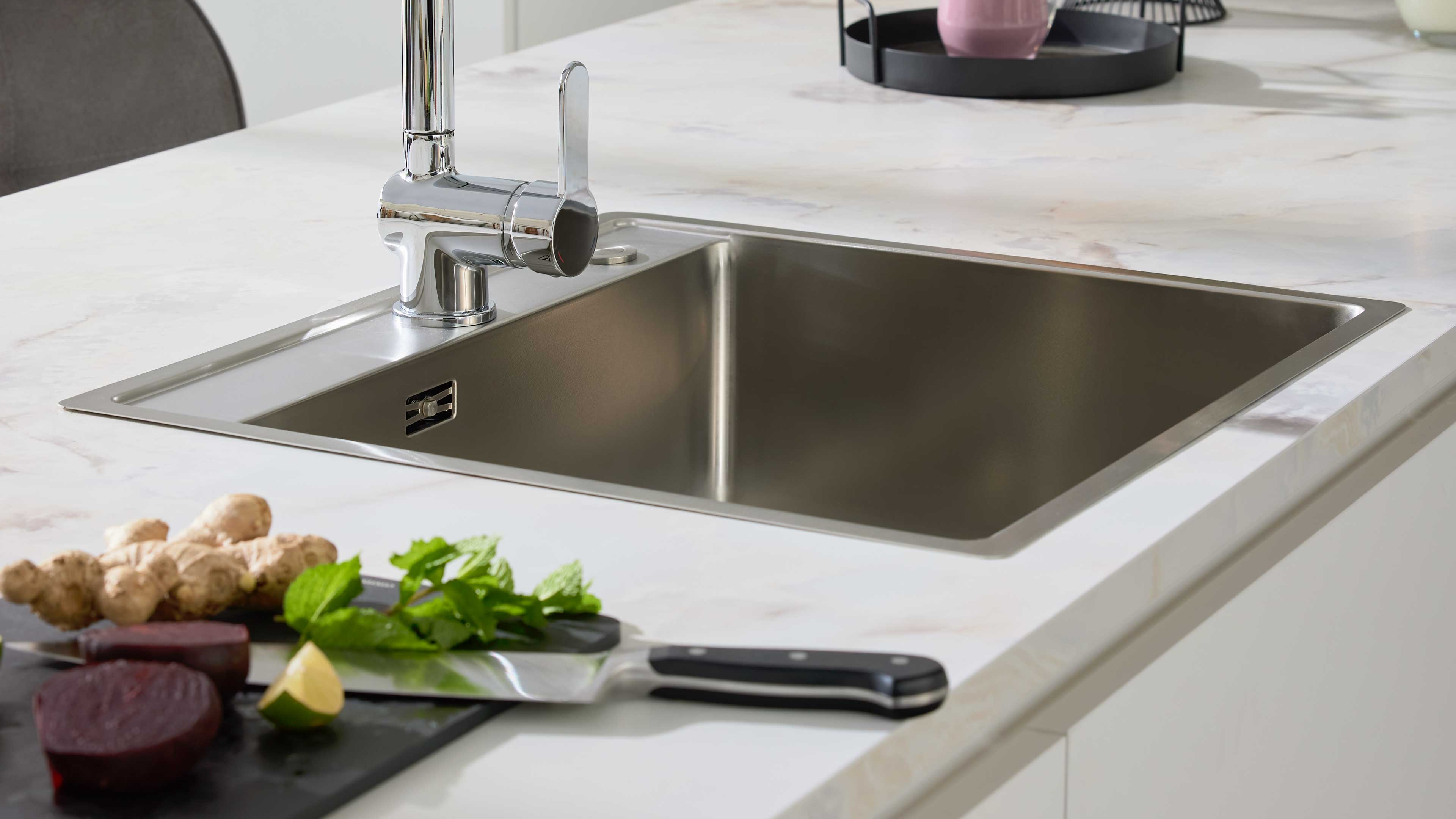 Edelstahl-Spüle mit verchromtem Einhebelmischer in weißer Arbeitsplatte, vorn Schneidebrett mit Messer, Rote Bete, Ingwer und Kräutern.