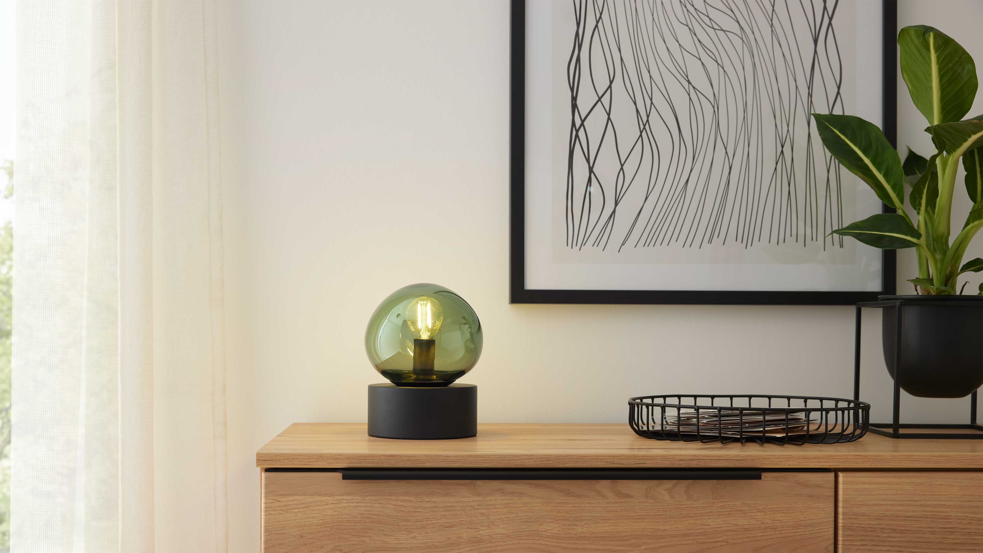 Tischlampe mit grüner Glaskugel auf Holzkommode, daneben schwarzes Drahtkorb-Tablett, rechts Topf mit Blättern und gerahmte Liniengrafik.