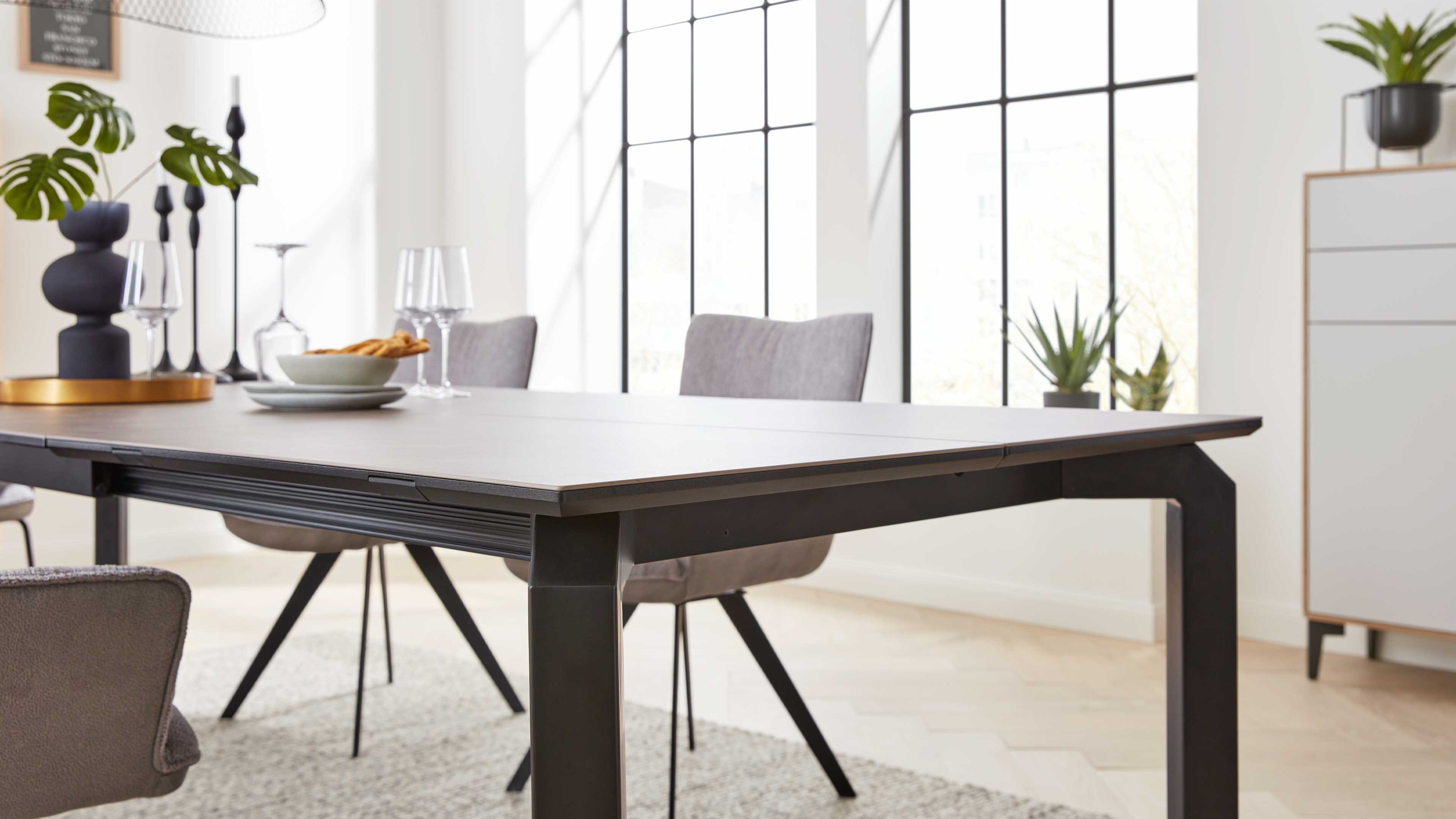 Rechteckiger Esstisch mit heller Holzplatte und schwarzem Metallgestell, umgeben von grauen Polsterstühlen, Gläsern und Pflanzen.