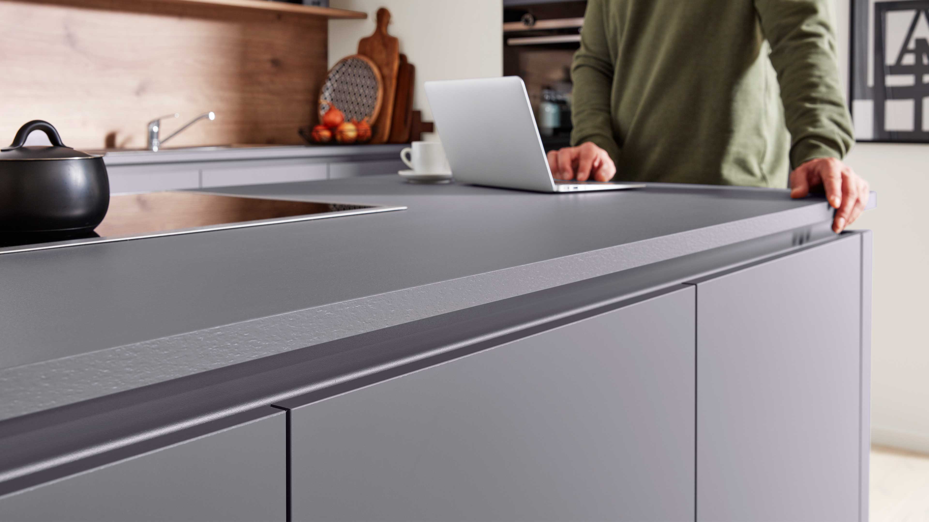 Graue Kücheninsel mit Griffkante, mattgrauer Arbeitsplatte, Induktionsfeld mit schwarzem Topf, Laptop und Person in grünem Pullover.