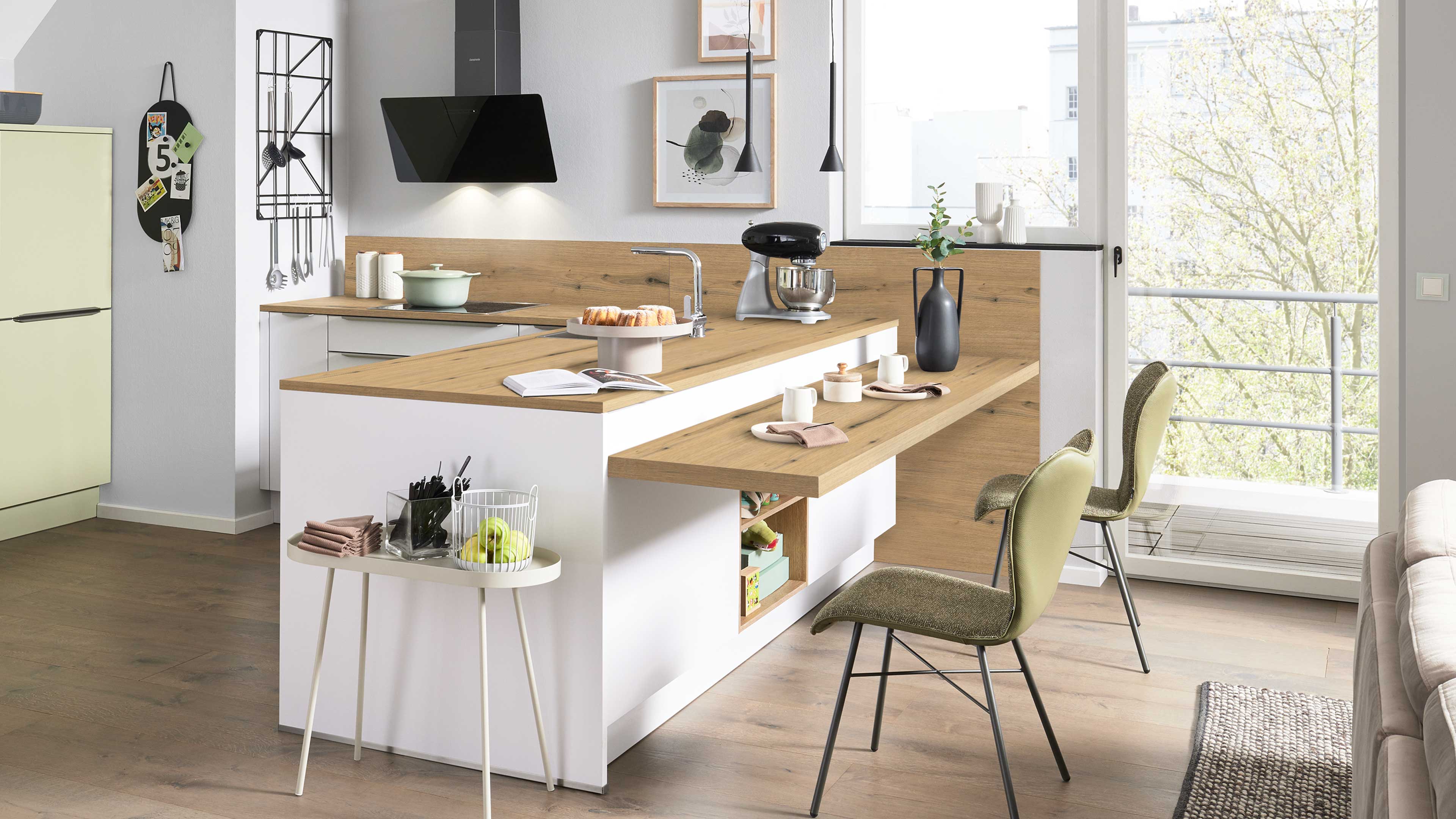 Kücheninsel mit Holzarbeitsplatte und Bartheke, zwei olivgrüne Stühle, Küchenmaschine, Vase und offenes Balkonfenster.