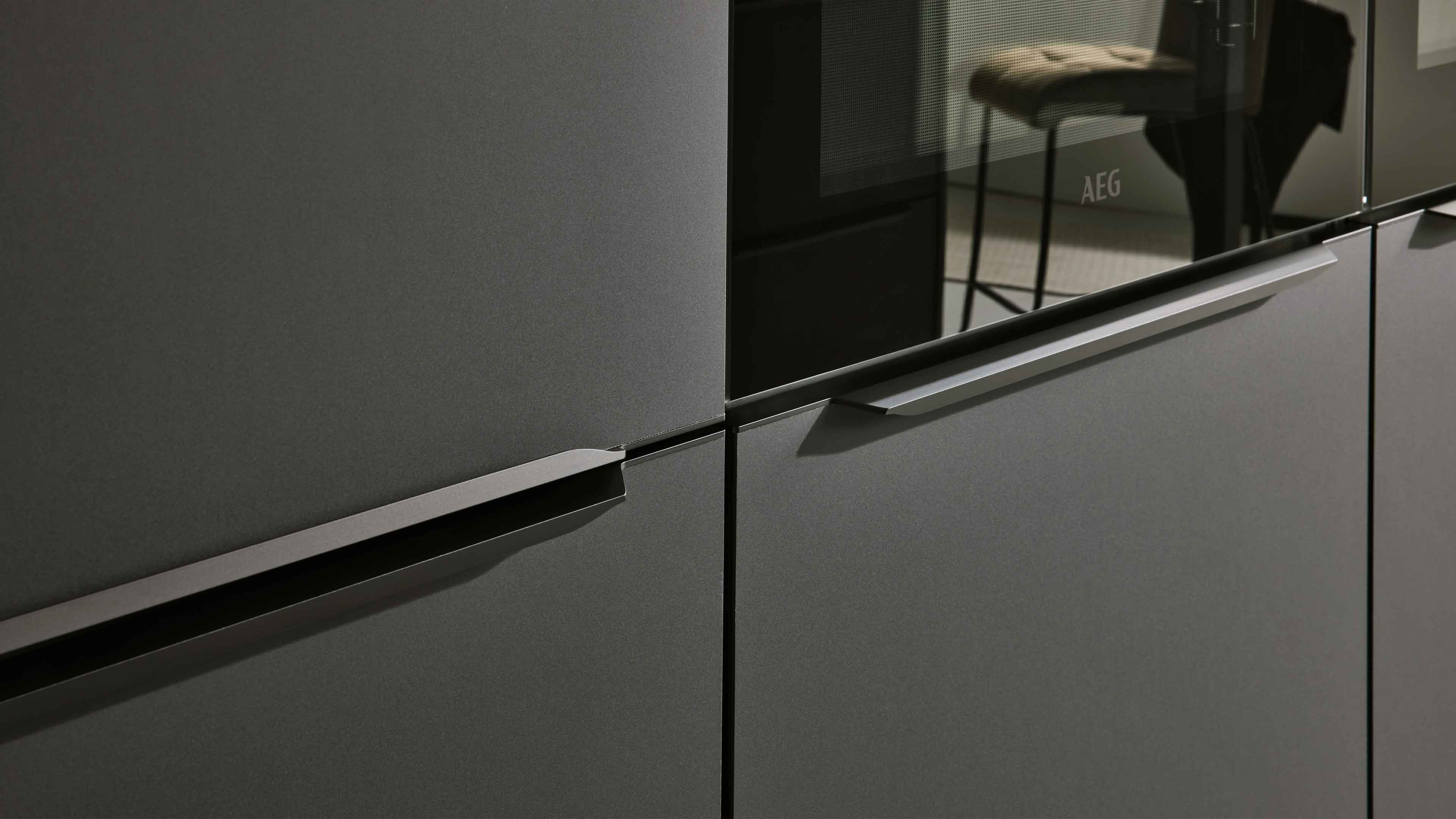 Grau matte Küchenschrankfronten mit eingelassenen horizontalen Griffleisten, rechts eine Glastür mit AEG-Logo.