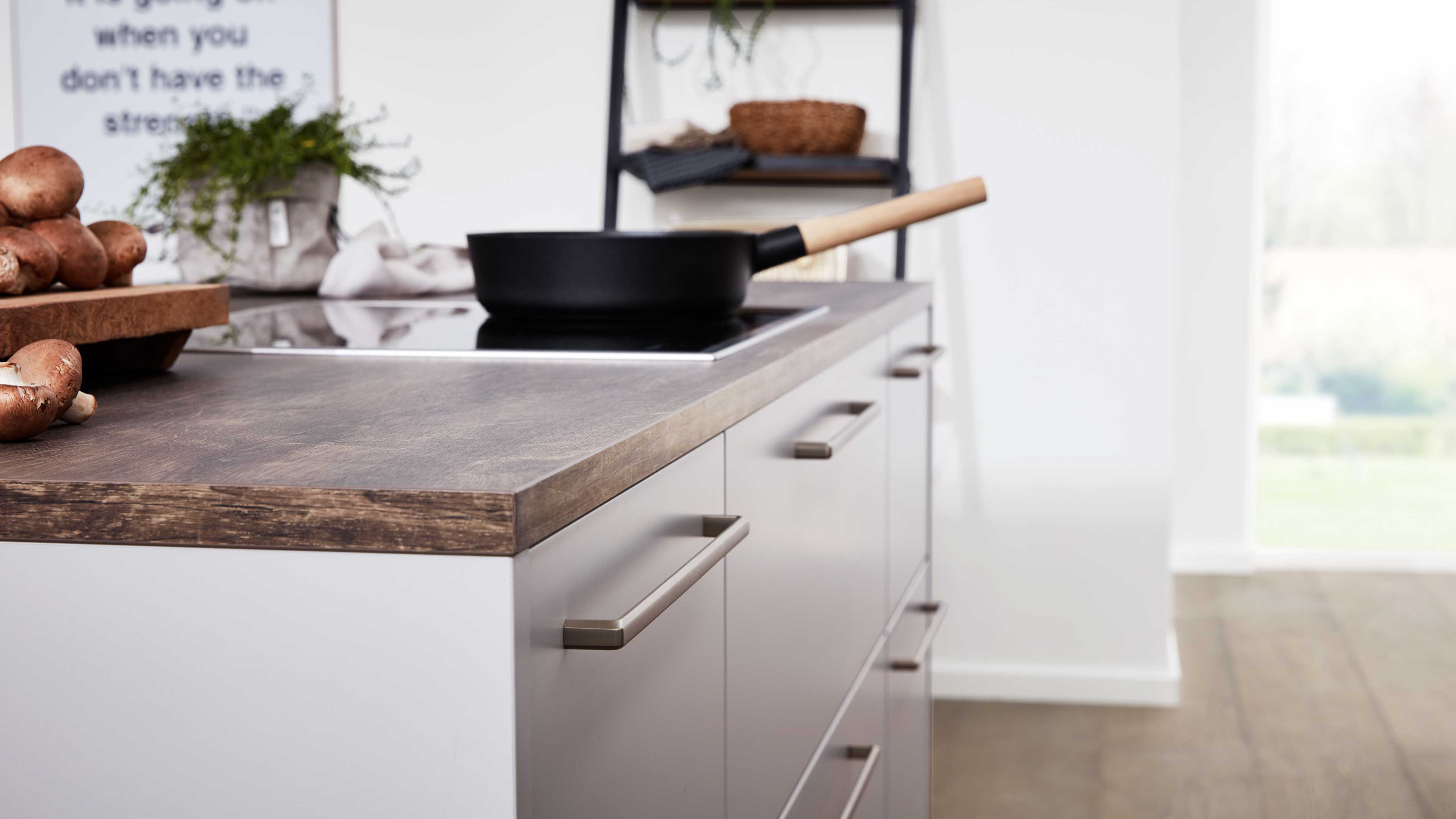 Kücheninsel mit dunkel gemaserter Holzarbeitsplatte, eingelassenem Kochfeld mit Pfanne und Metallgriffleisten an Schubladen.