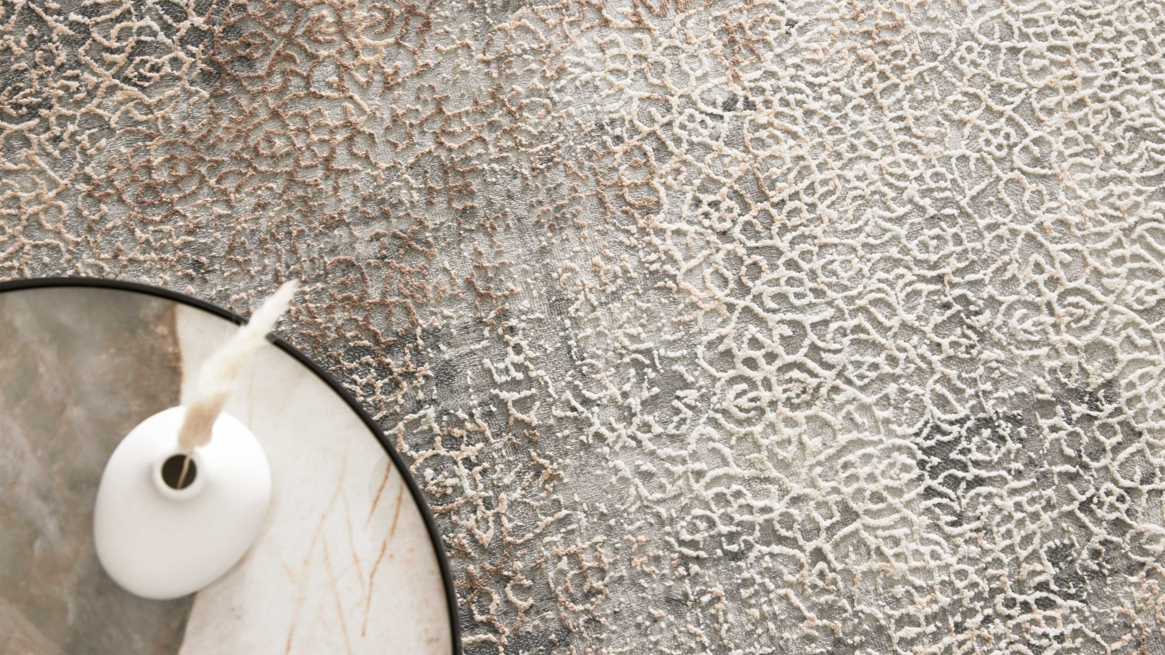 Grau-beigefarbener Teppich mit erhabenem, ornamentalem Muster, links ein runder Marmortisch mit weißer Keramikvase und trockenem Pampasgras.