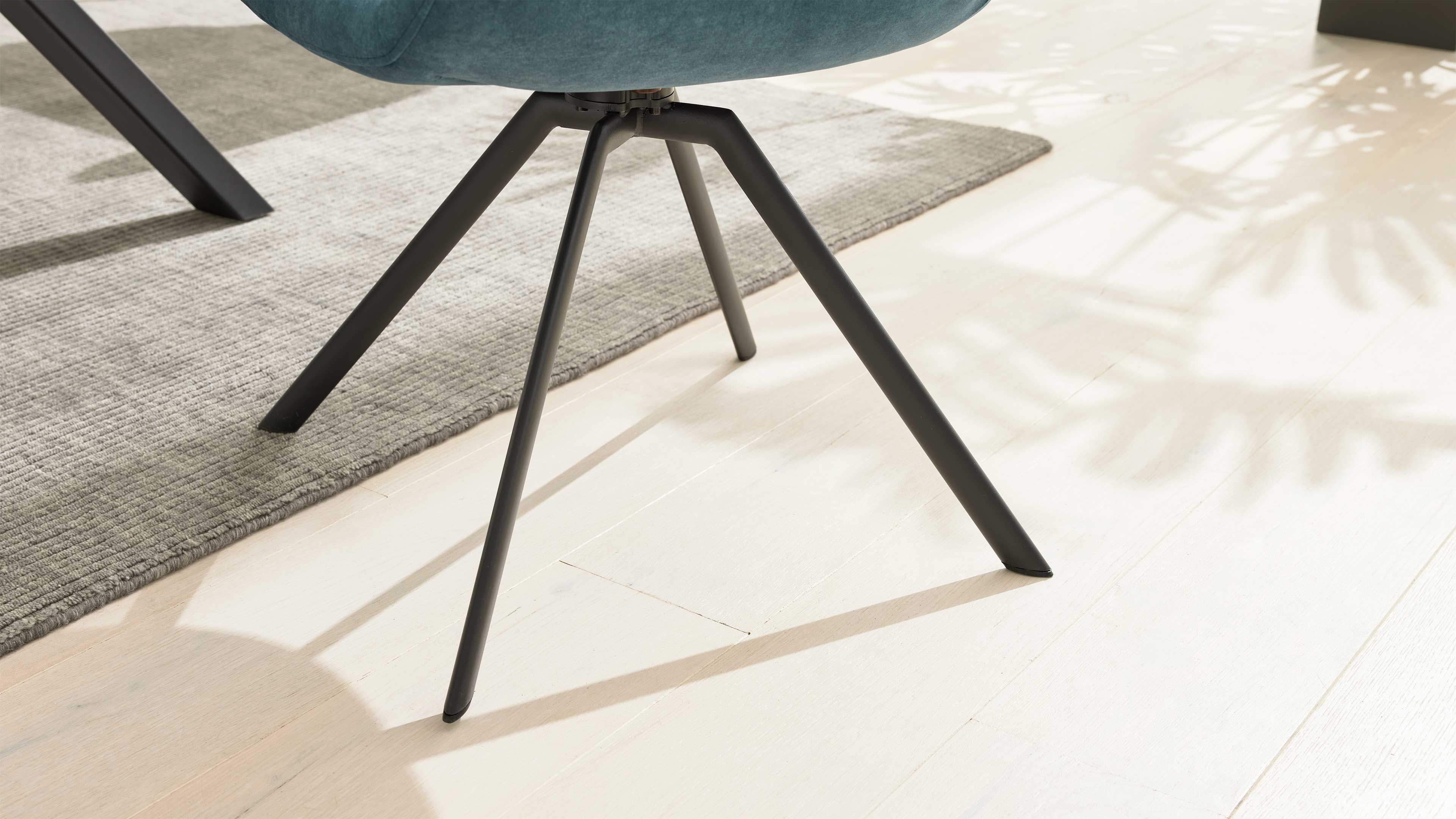 Schwarze, schrägstehende Metallbeine eines Stuhls unter blaugrünem Sitz auf hellem Holzboden, neben grauem Teppich und Pflanzenschatten.