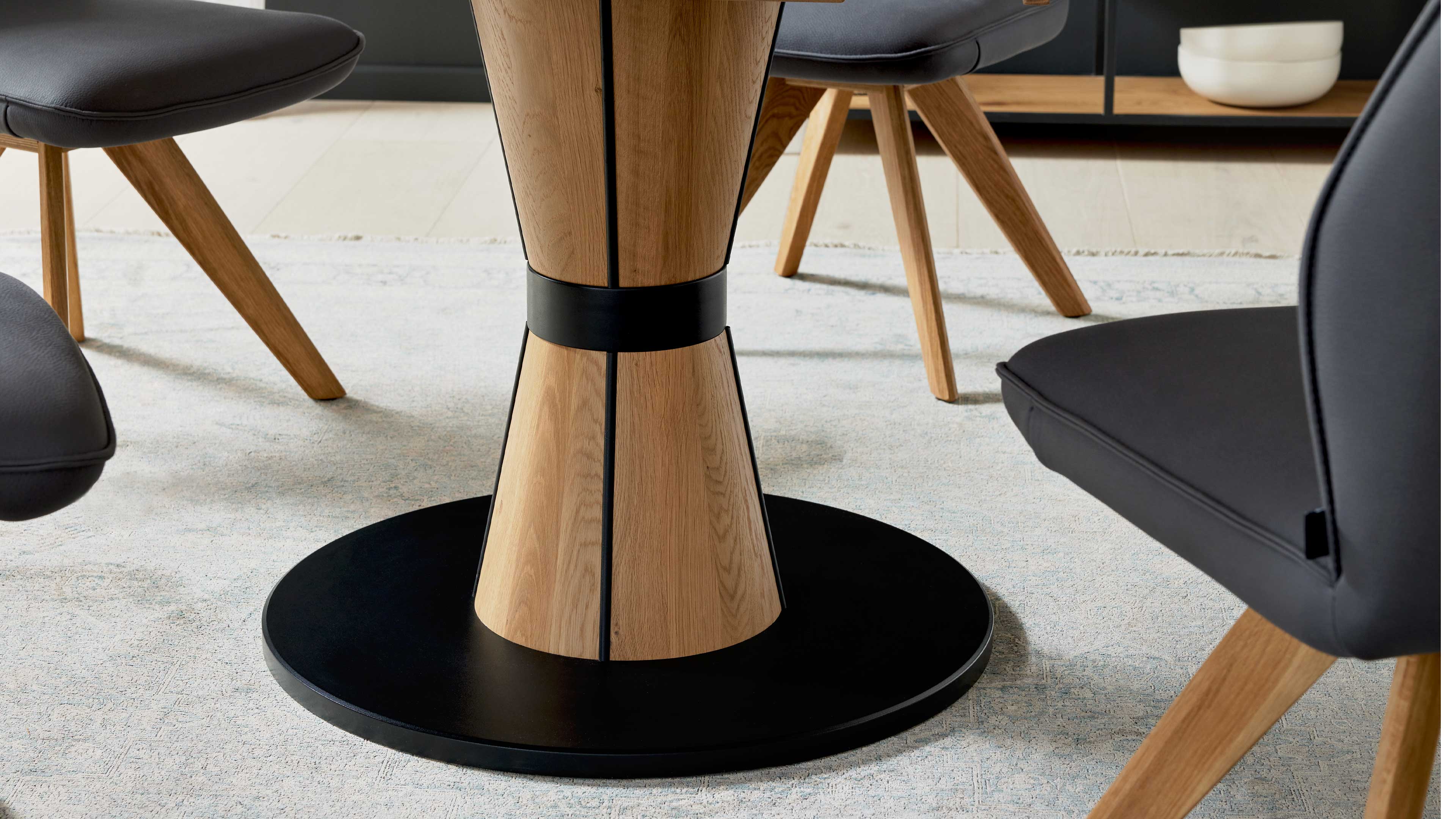 Segmentierter Holz-Tischfuß mit schwarzem Metallring und rundem schwarzem Sockel, dazu schwarze gepolsterte Stuhlsitze auf Holzbeinen.