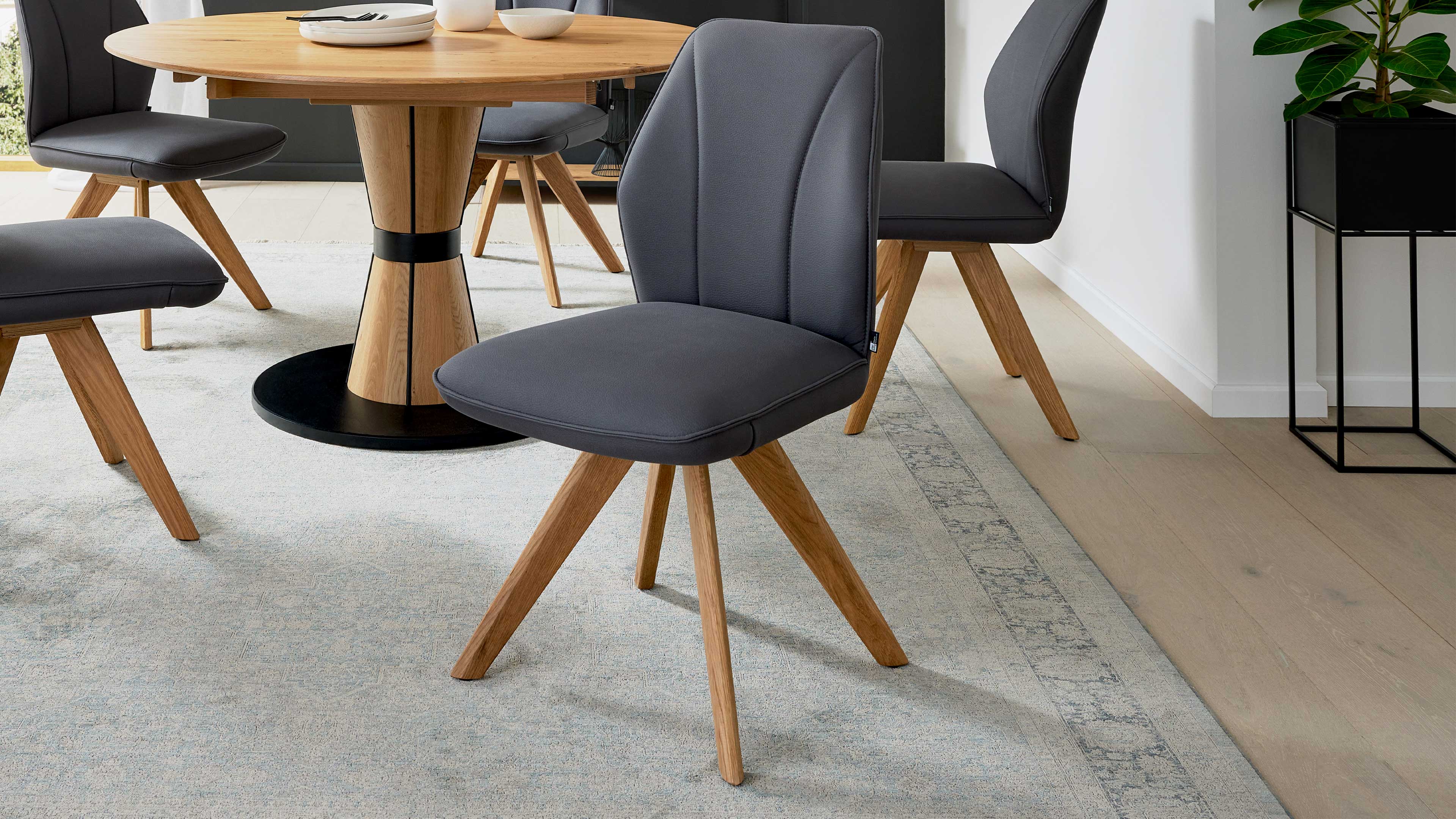Grau gepolsterter Esszimmerstuhl mit geschwungenem Rücken und vier schrägen Eichenholzbeinen vor rundem Holztisch und Teppich.