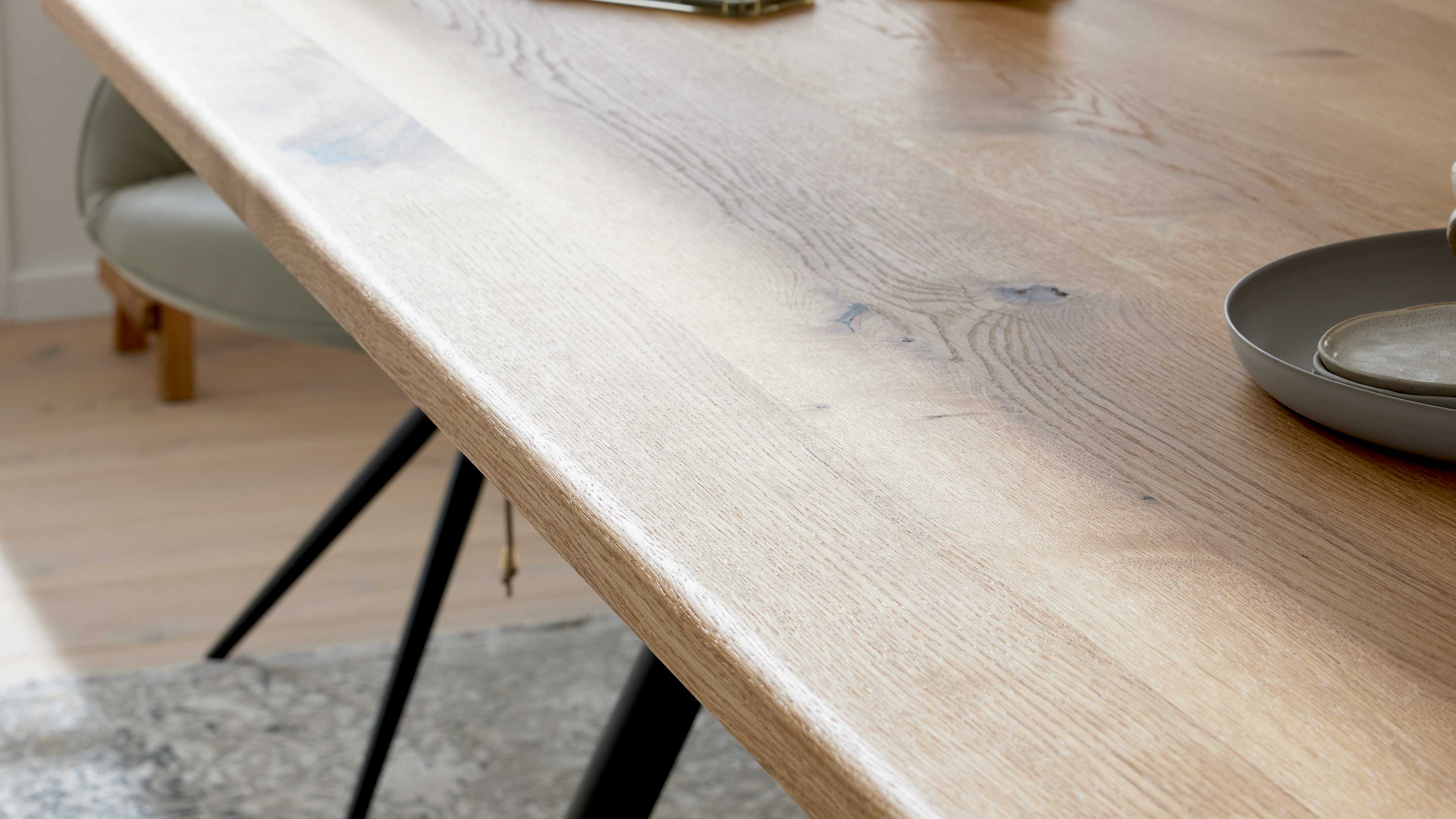 Helle Tischplatte mit sichtbarer Holzmaserung und abgerundeter Kante, schwarze Metallbeine, graue Schale und Stuhl im Hintergrund.
