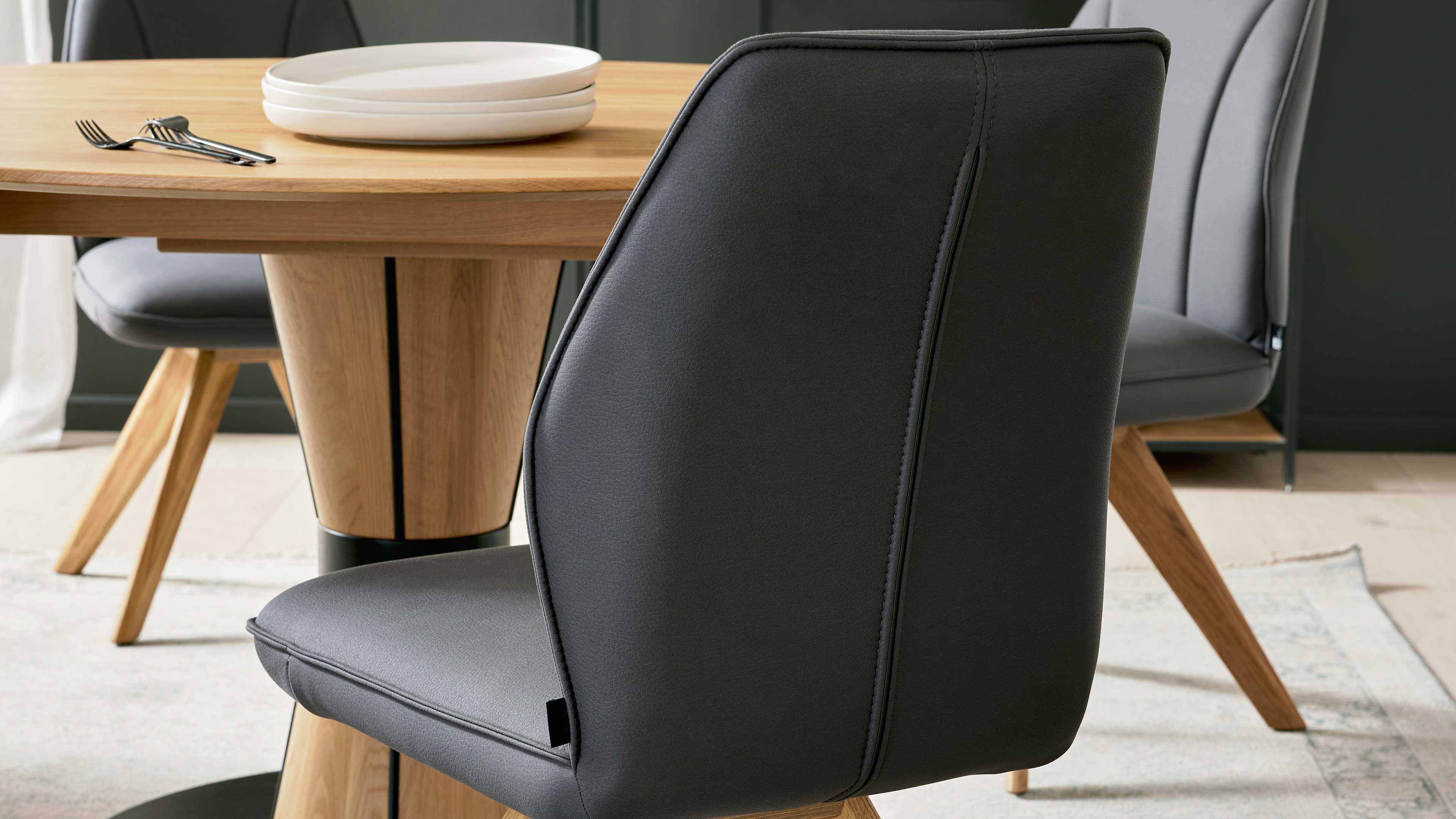 Dunkelgrauer, gepolsterter Esszimmerstuhl im Vordergrund, dahinter runder Esstisch aus hellem Holz mit gestapelten Tellern und Besteck.