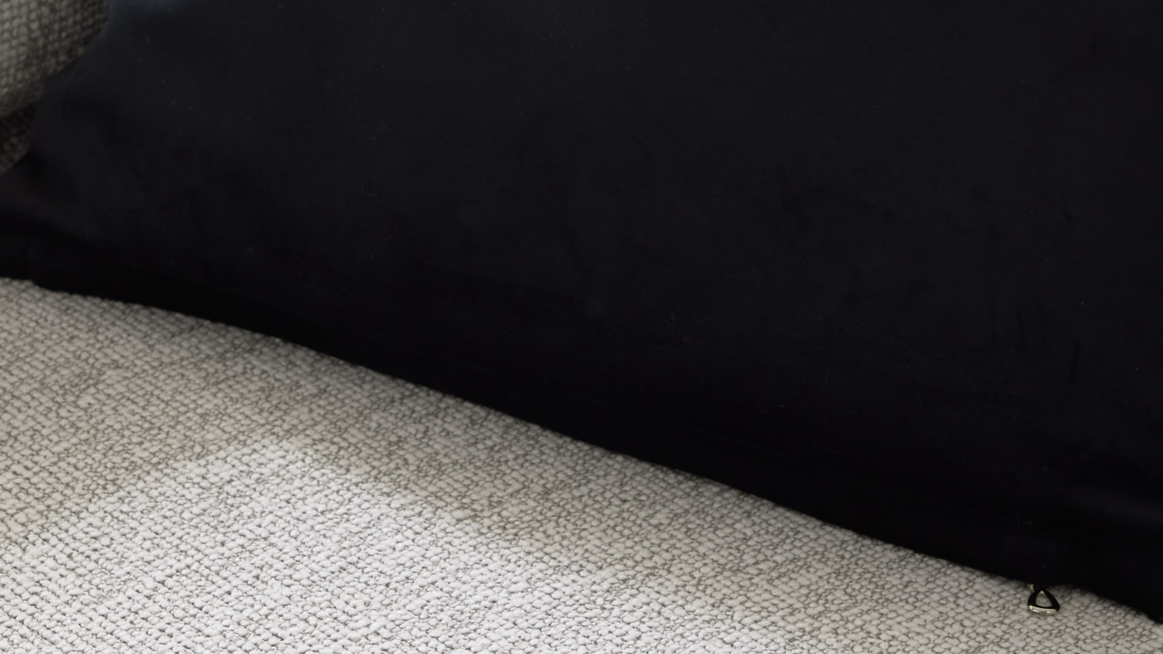Schwarzes Kissen auf hellgrauem, grob gewebtem Sofa, mit sichtbarer Nahtkante und kleinem metallischem Reißverschlussanhänger am Rand.