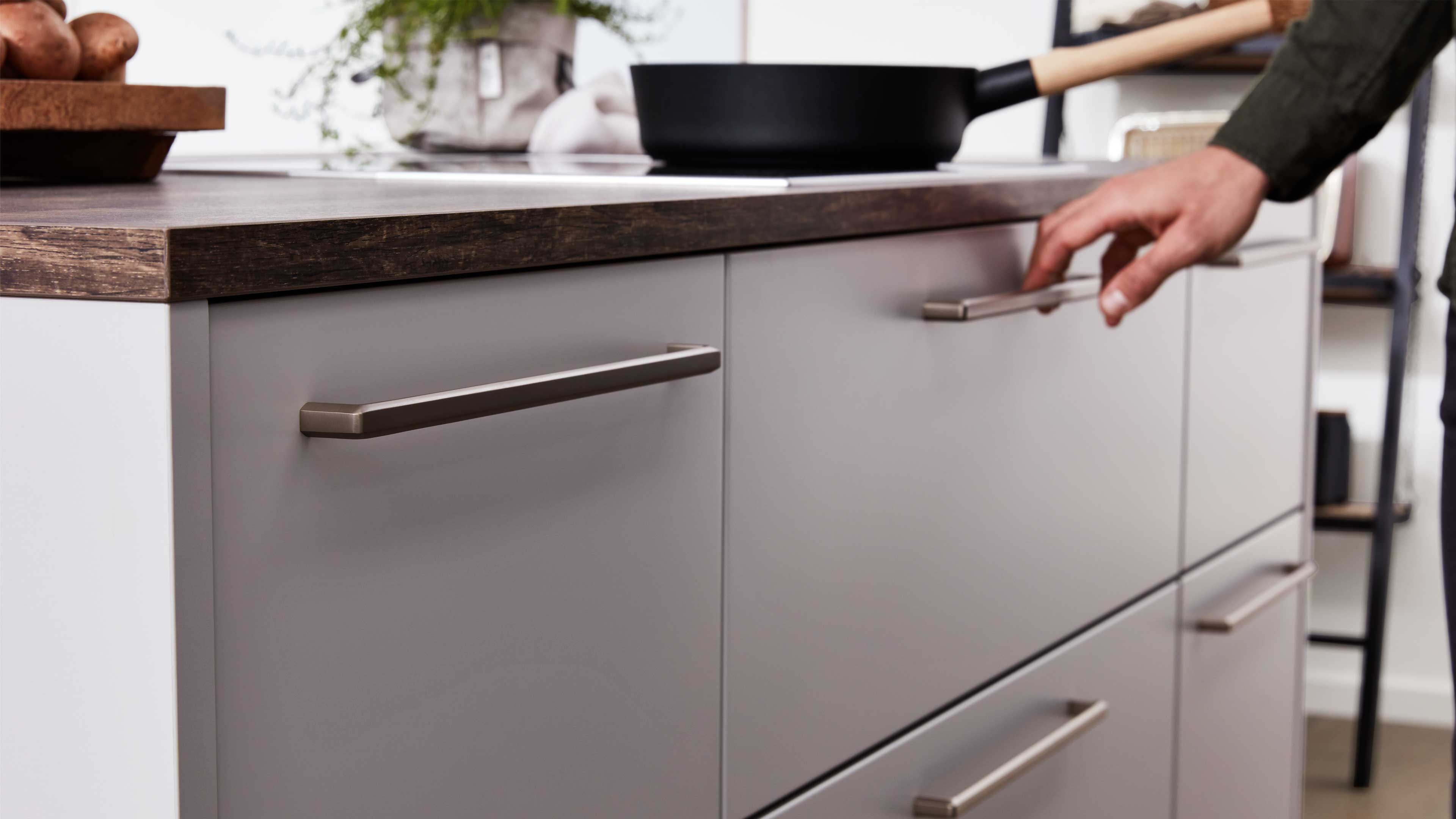 Grau matte Küchenschublade mit langen Metallgriffen, dunkler Holzarbeitsplatte, Pfanne und Hand am Griff.