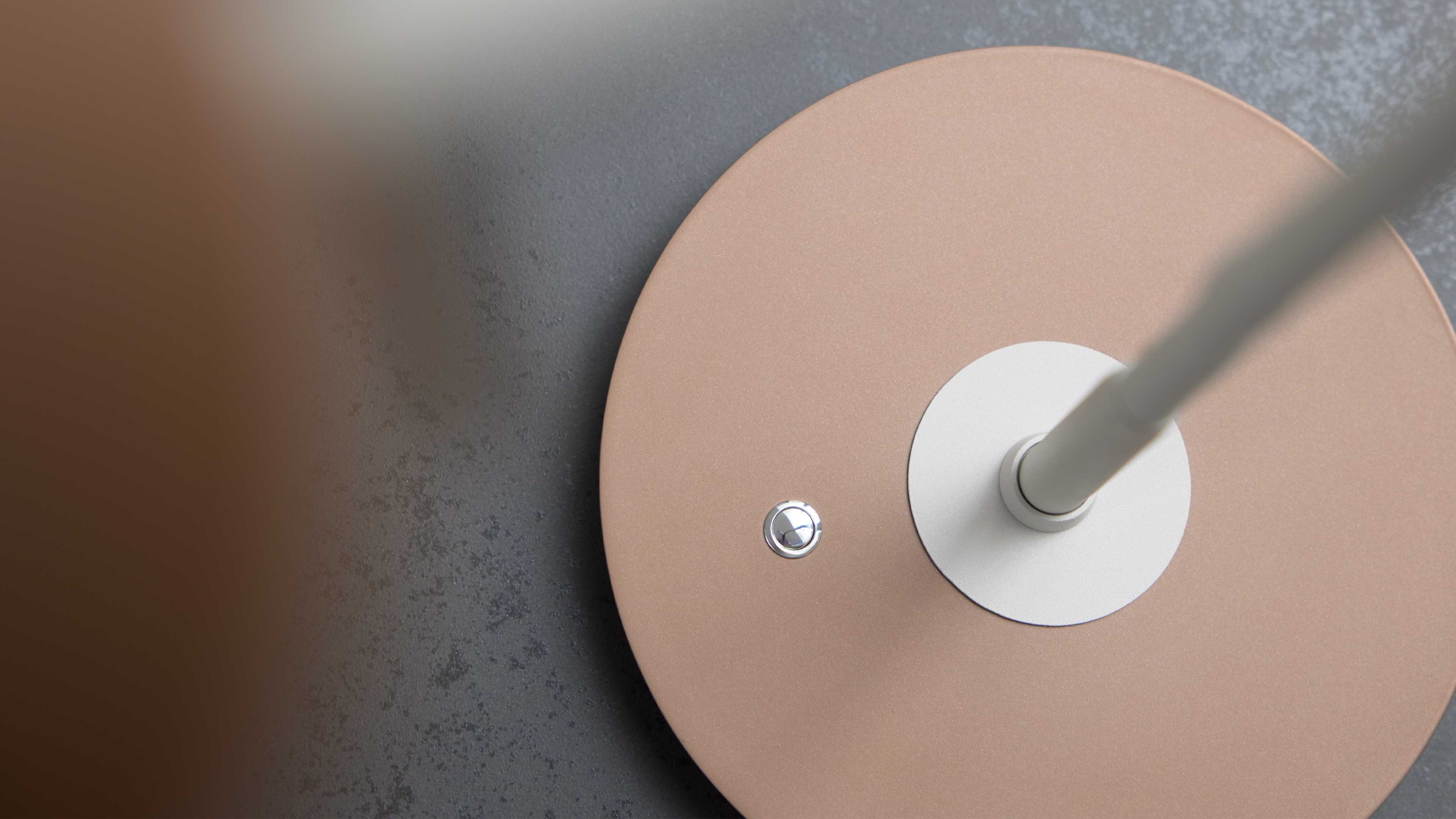 Runder Lampenfuß in Roségold mit weißem Befestigungsring und kleinem silbernem Druckknopf auf strukturierter grauer Oberfläche.