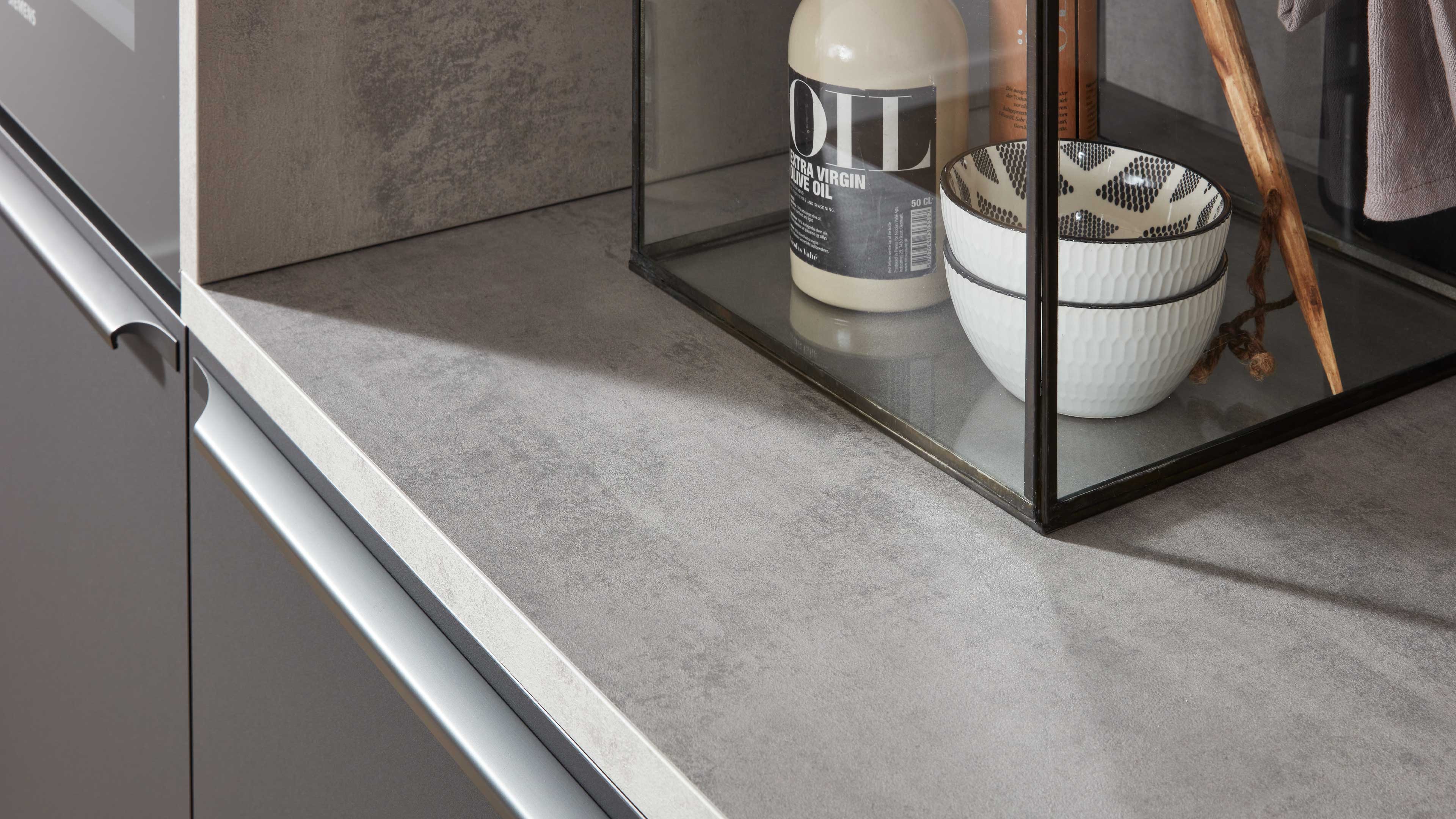 Graue Küchenarbeitsplatte mit hellerer Kante, darauf ein schwarzer Glasbehälter mit zwei weiß gemusterten Schalen und einer Ölflasche.