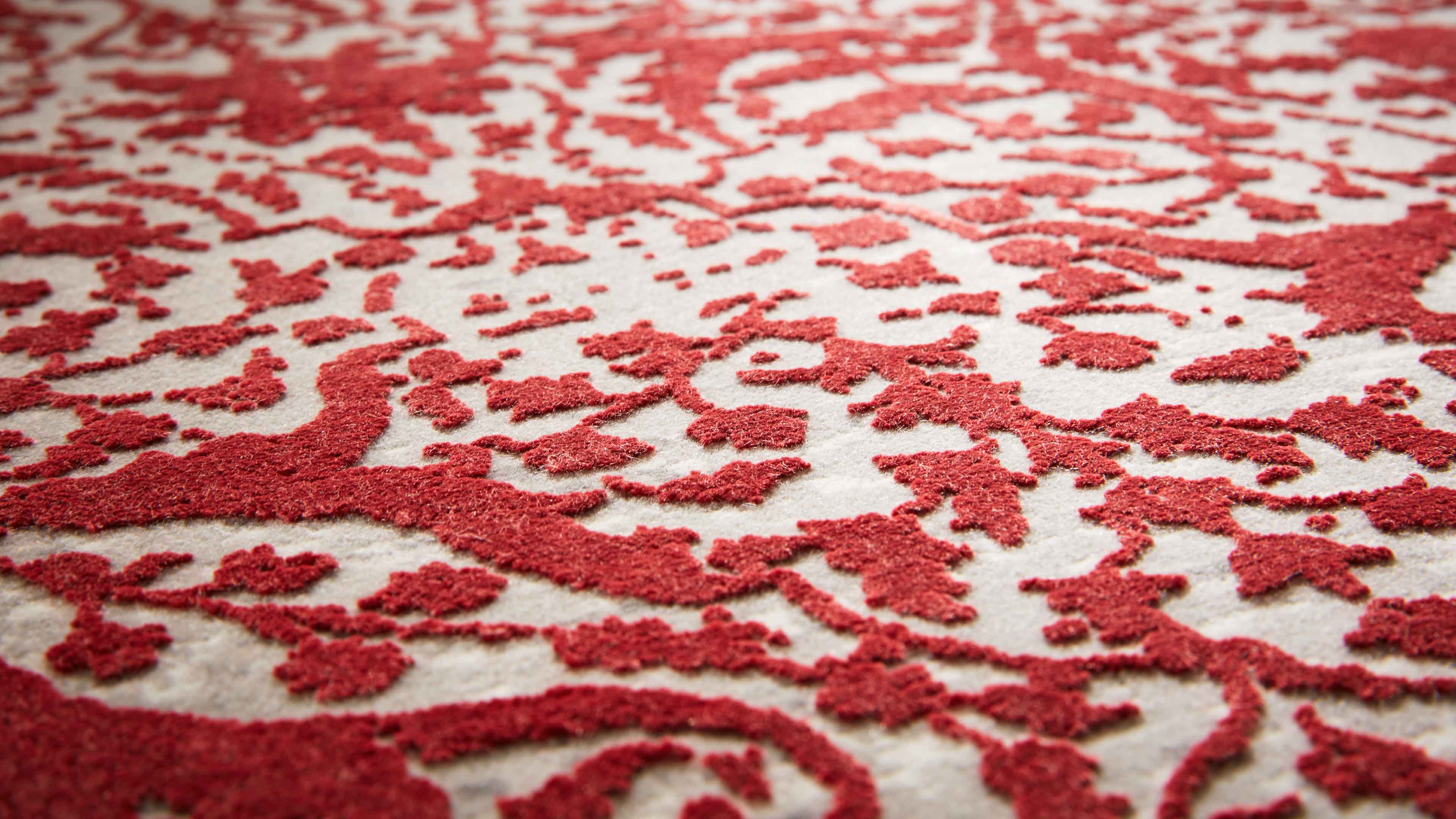 Nahaufnahme eines Teppichs mit erhabenen roten floralen Ornamenten auf hellbeigem Grund, grobe unregelmäßige Florstruktur sichtbar.