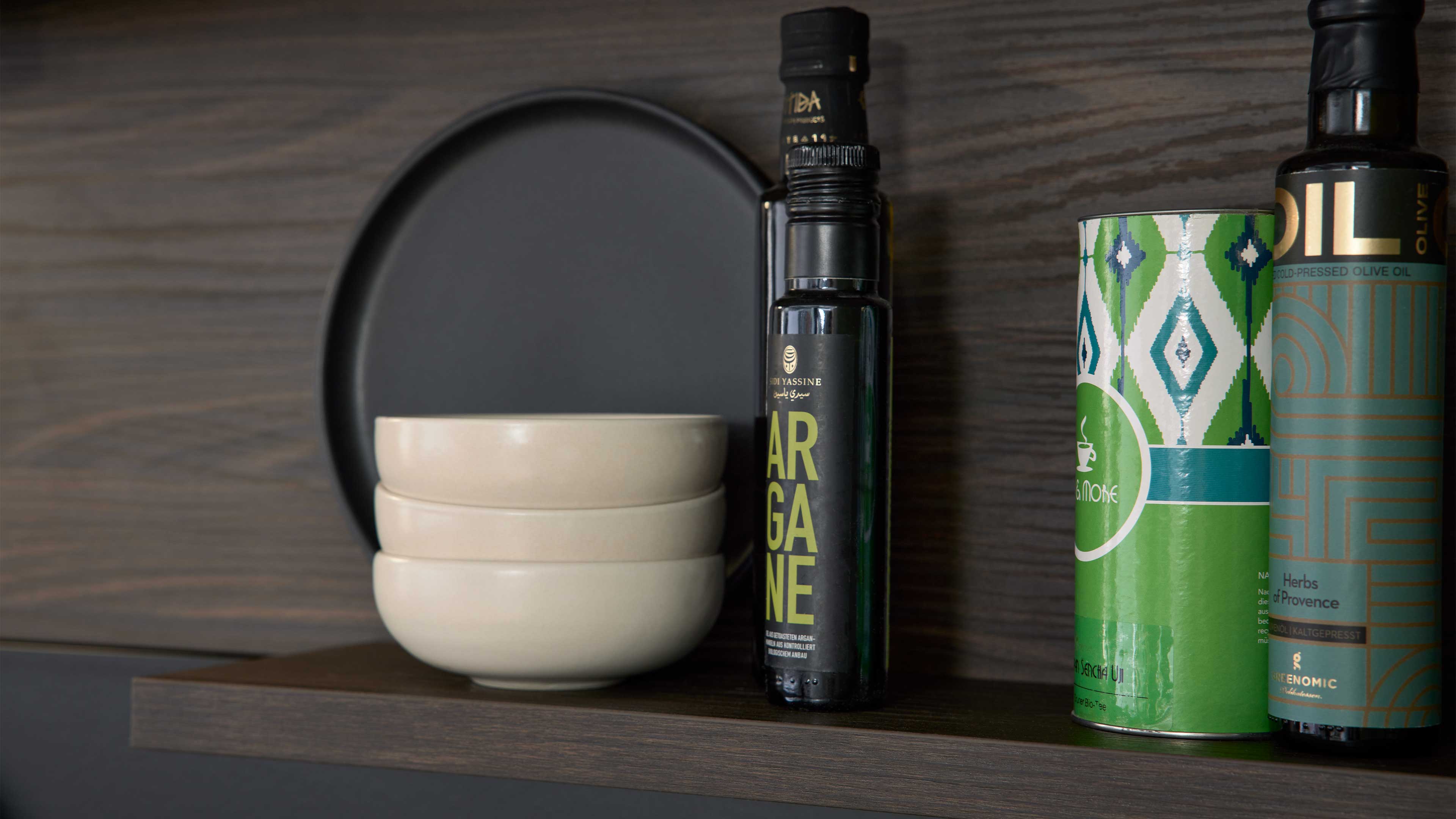 Drei kleine beige Schalen übereinander auf dunklem Holzregal vor einem schwarzen Teller, daneben Ölflaschen und Blechdose.