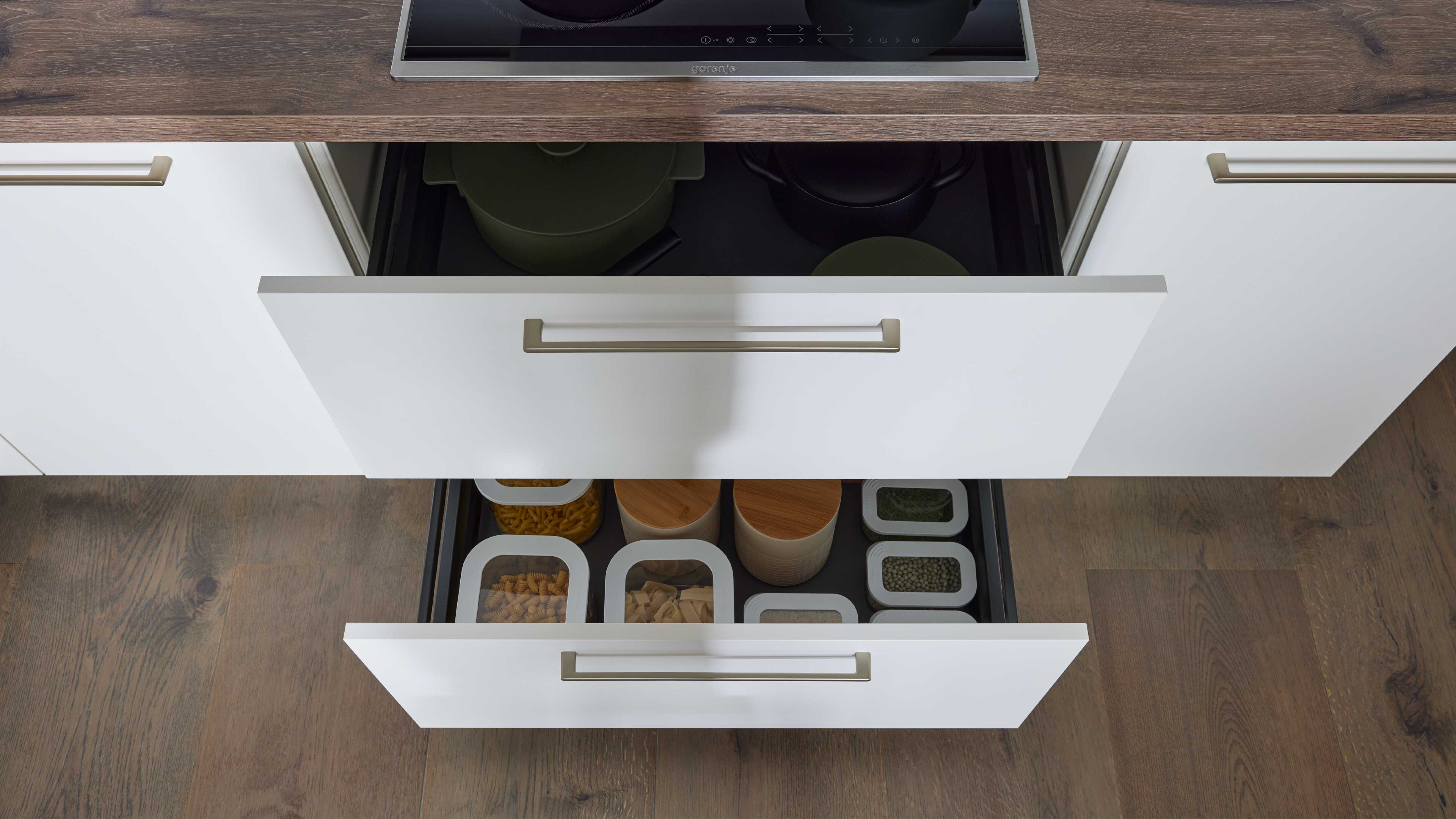 Zwei geöffnete weiße Küchenschubladen unter einer Holzarbeitsplatte: oben Töpfe, unten sortierte Vorratsbehälter mit Nudeln und Hülsenfrüchten.