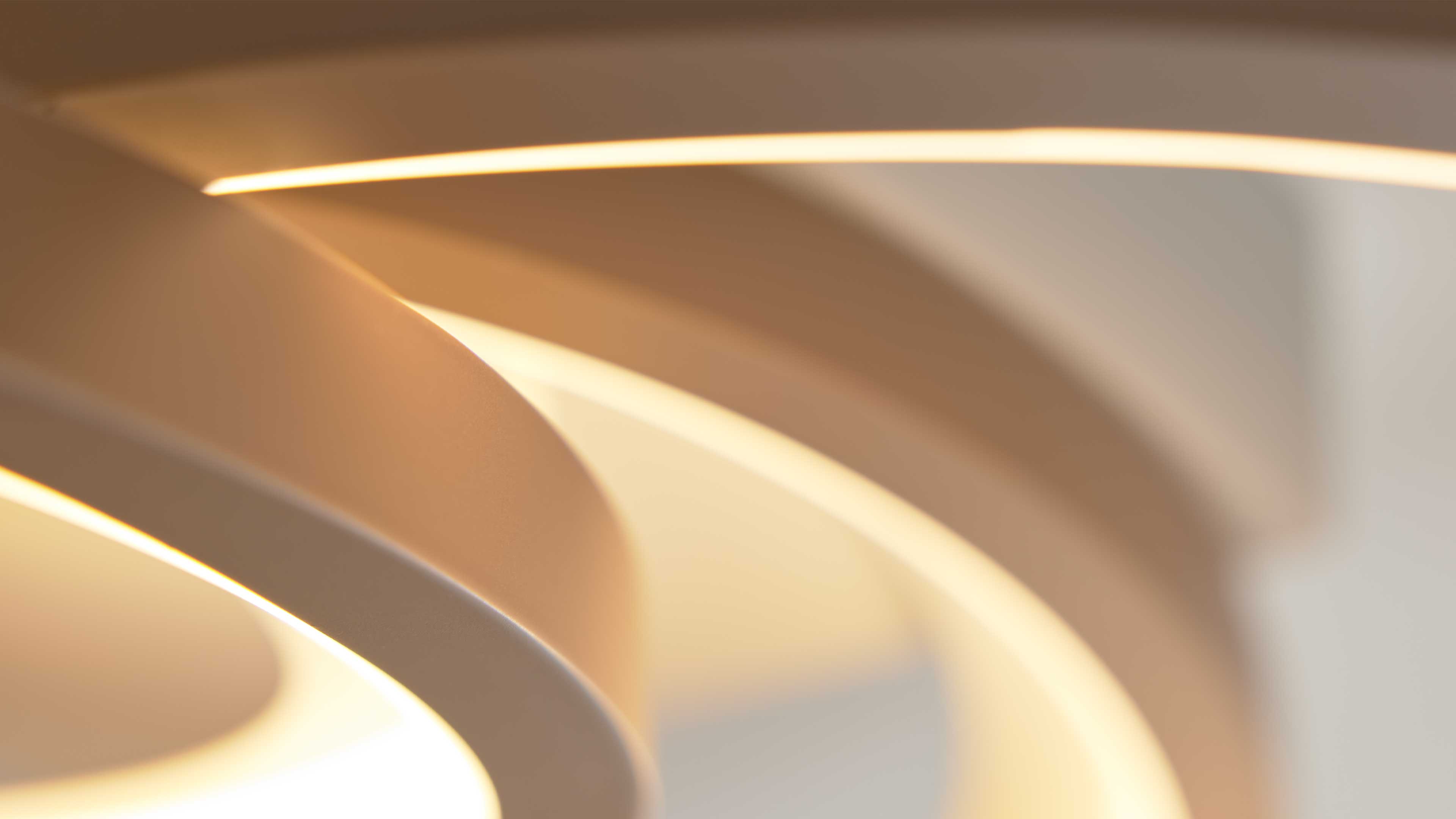 Rund geschwungene, übereinanderliegende Ringe einer Leuchte in cremefarbenem Ton, die warmweißes Licht abstrahlen und weiche Schatten bilden.