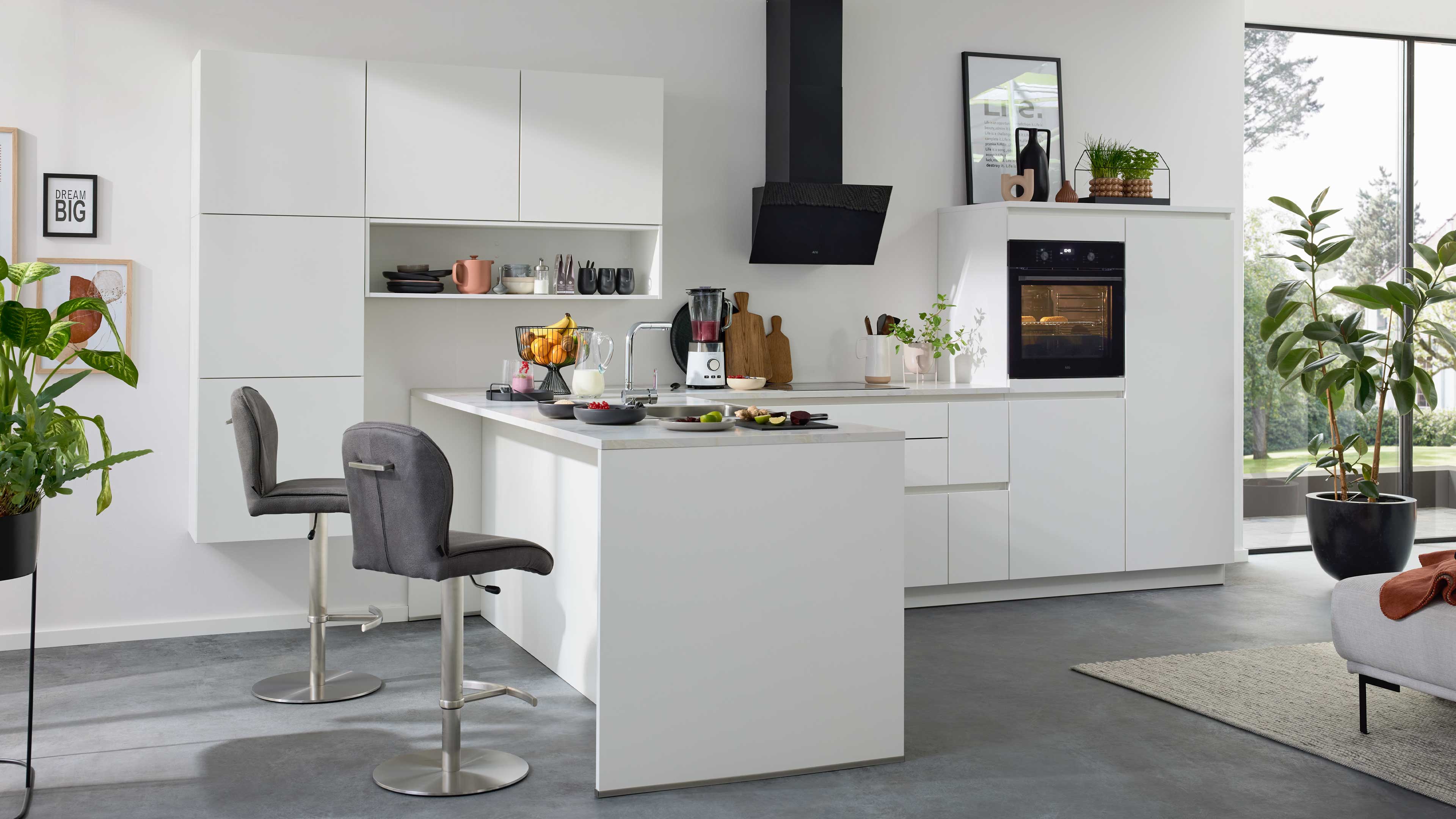 Weiße Einbauküche mit Insel und zwei grauen Barhockern, schwarzer Dunstabzugshaube, Ofen und Pflanzen am Fenster.