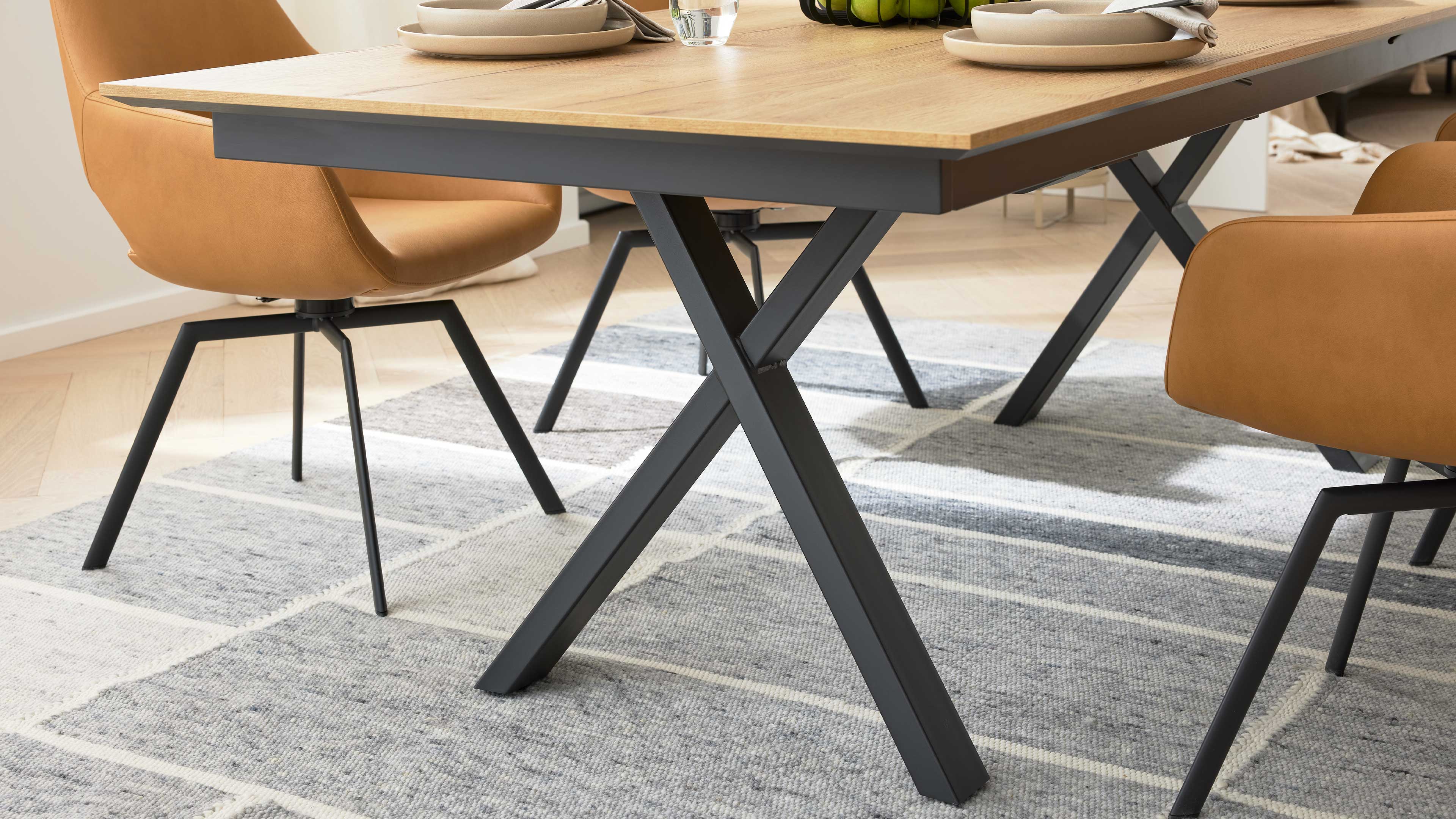 Esstisch mit heller Holzplatte und schwarzen X-förmigen Metallbeinen, umgeben von braunen Stühlen auf grauem Teppich.