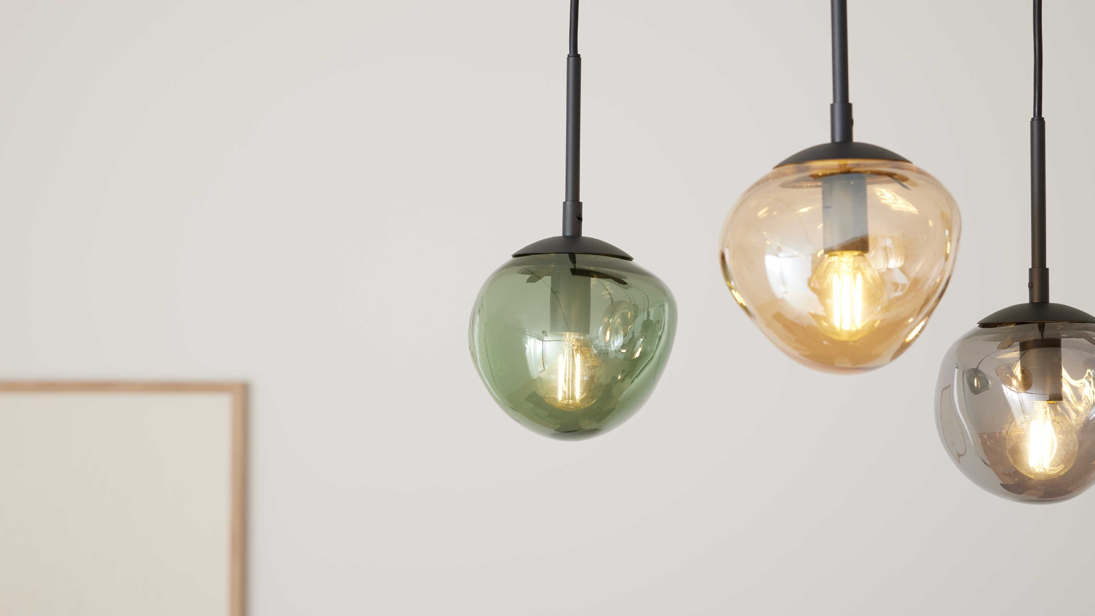 Grüne Glas-Pendelleuchte links mit sichtbarem Filament, daneben bernstein- und rauchgraue Lampen an schwarzen Kabeln vor heller Wand.