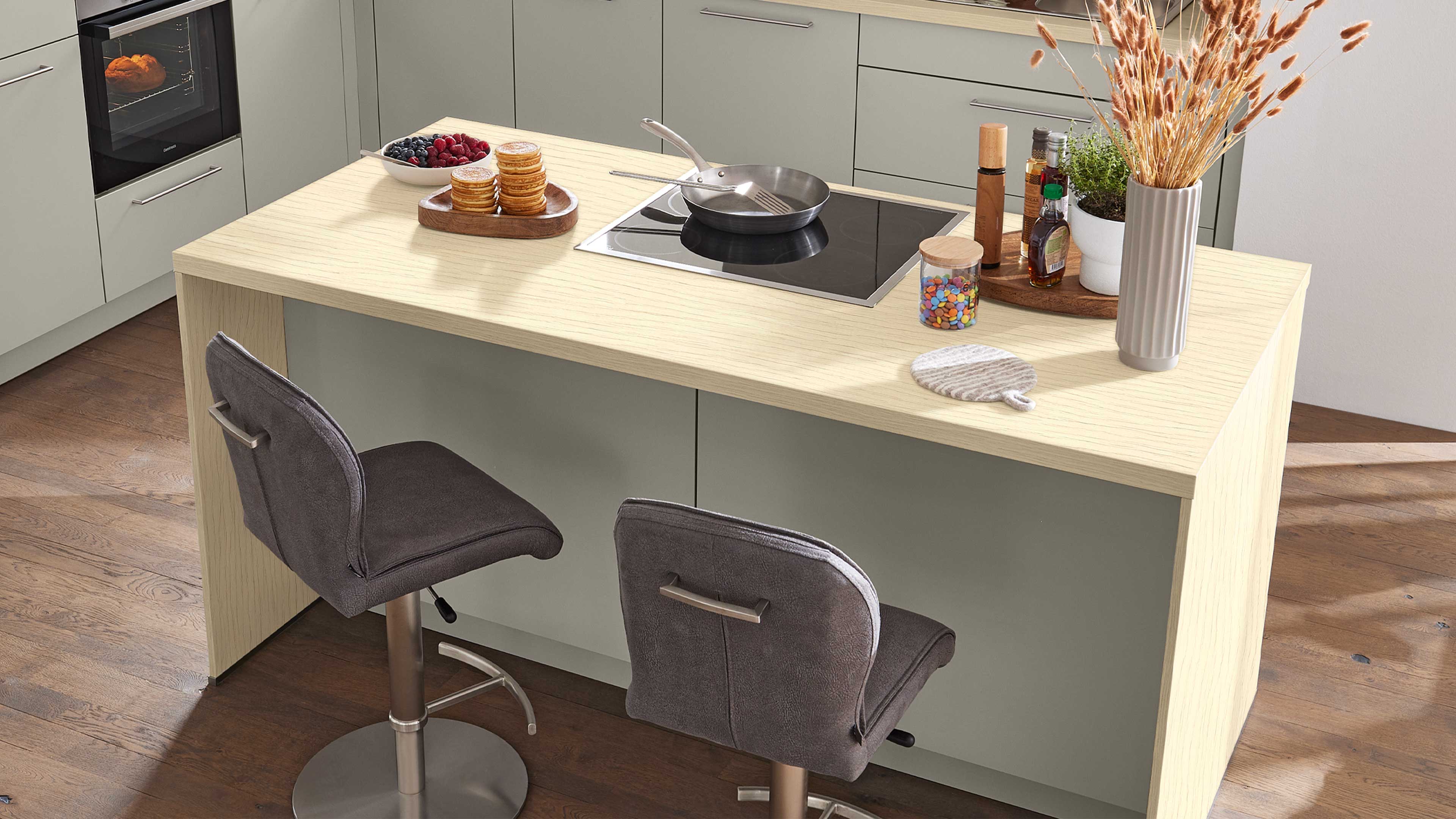 Kücheninsel mit heller Holzarbeitsplatte, schwarzem Kochfeld mit Pfanne, zwei grauen Barhockern und einer Vase mit getrockneten Halmen.