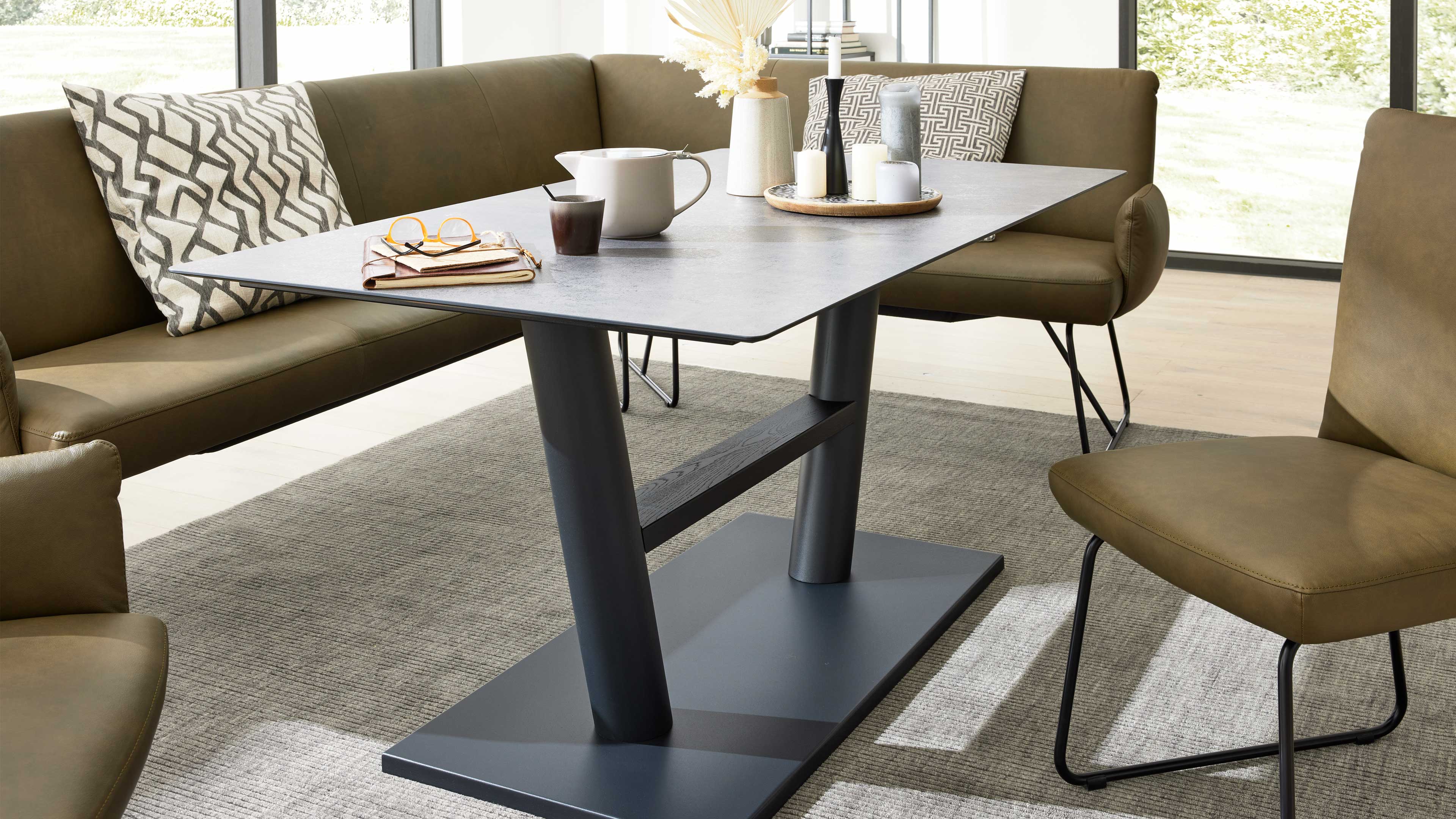 Rechteckiger grauer Tisch mit zwei schrägen Beinen, bestückt mit Kanne, Tasse und Kerzentablett, umgeben von olivgrünen Sitzmöbeln.