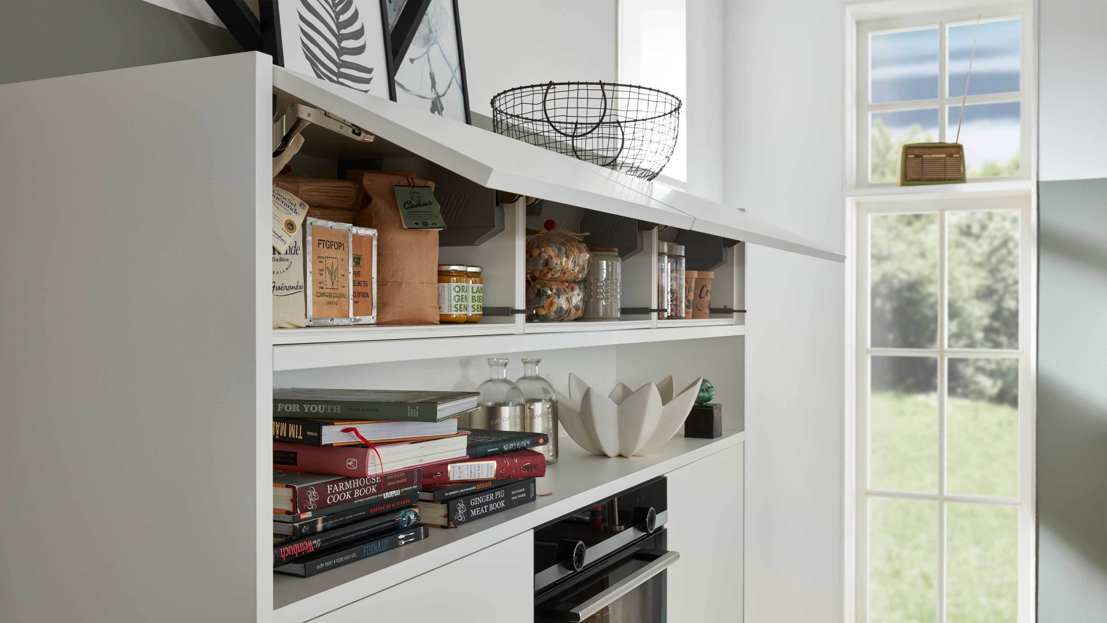 Weiße Küchenzeile mit aufgeklappter Front, offenem Fach mit Vorratsdosen, Papiertüten und Kochbüchern sowie Drahtkorb oben.