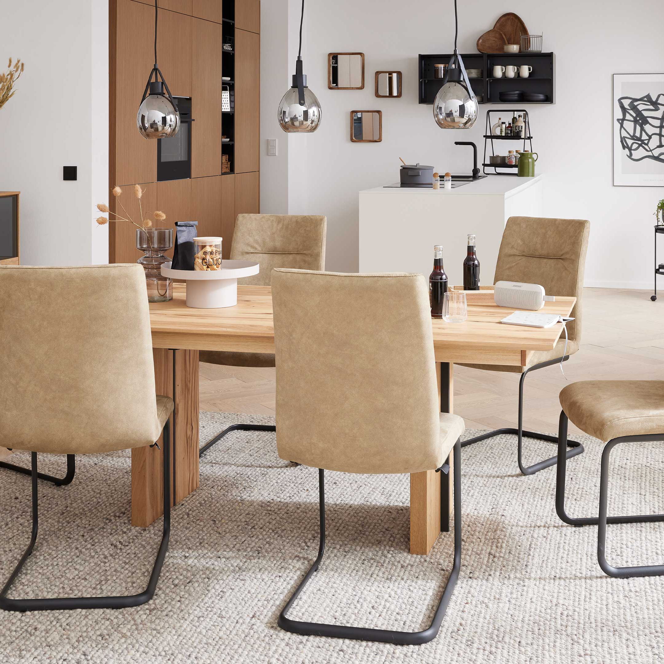 Rechteckiger Holztisch mit sechs beige gepolsterten Freischwinger-Stühlen, darauf Deko, vor Sideboard und Hängelampen.