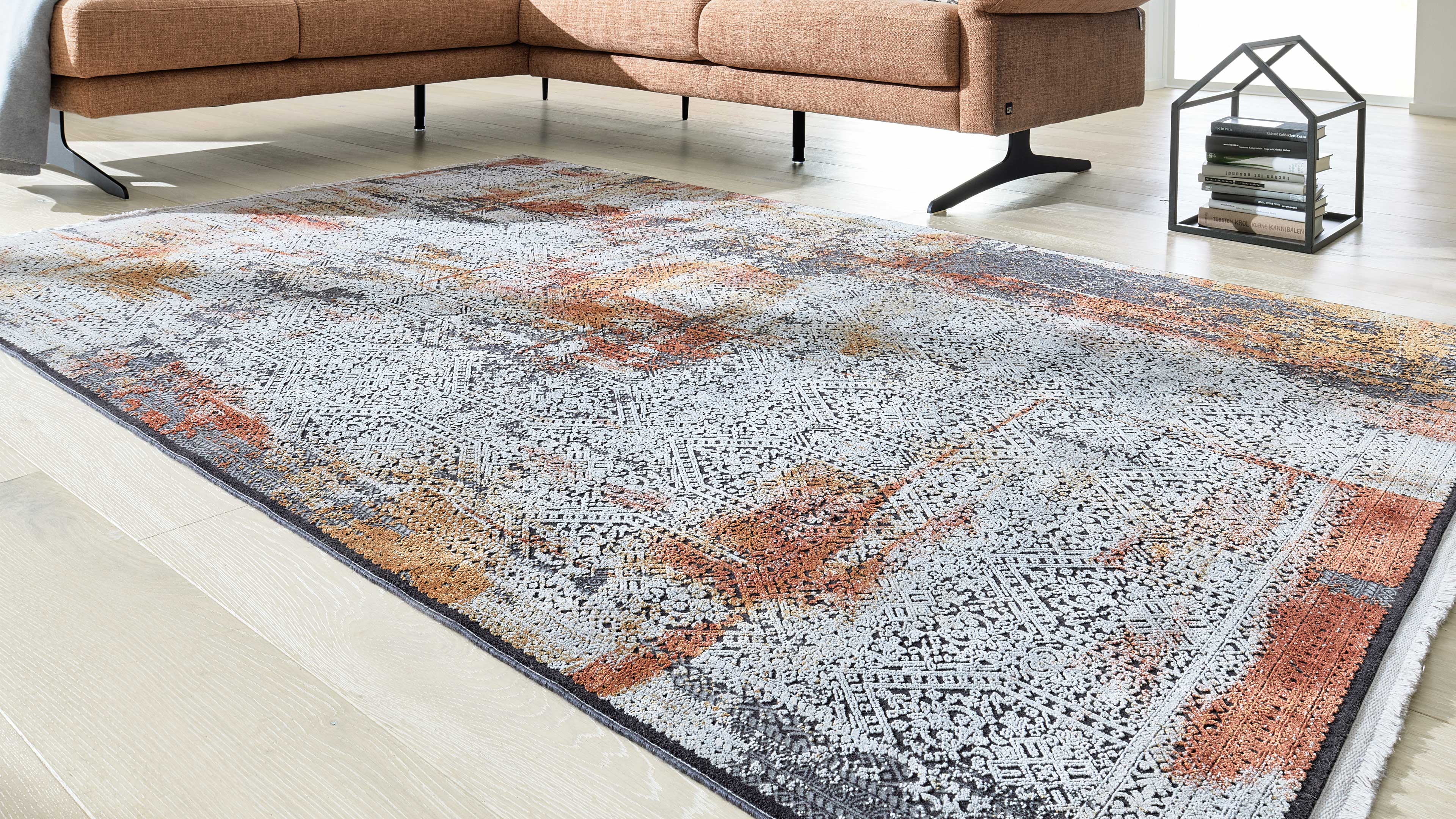 Großer Teppich mit weiß-grauem Ornamentmuster und rostfarbenen Sprenkeln auf hellem Holzfußboden, links Sofa, rechts offenes Metallregal mit Büchern.