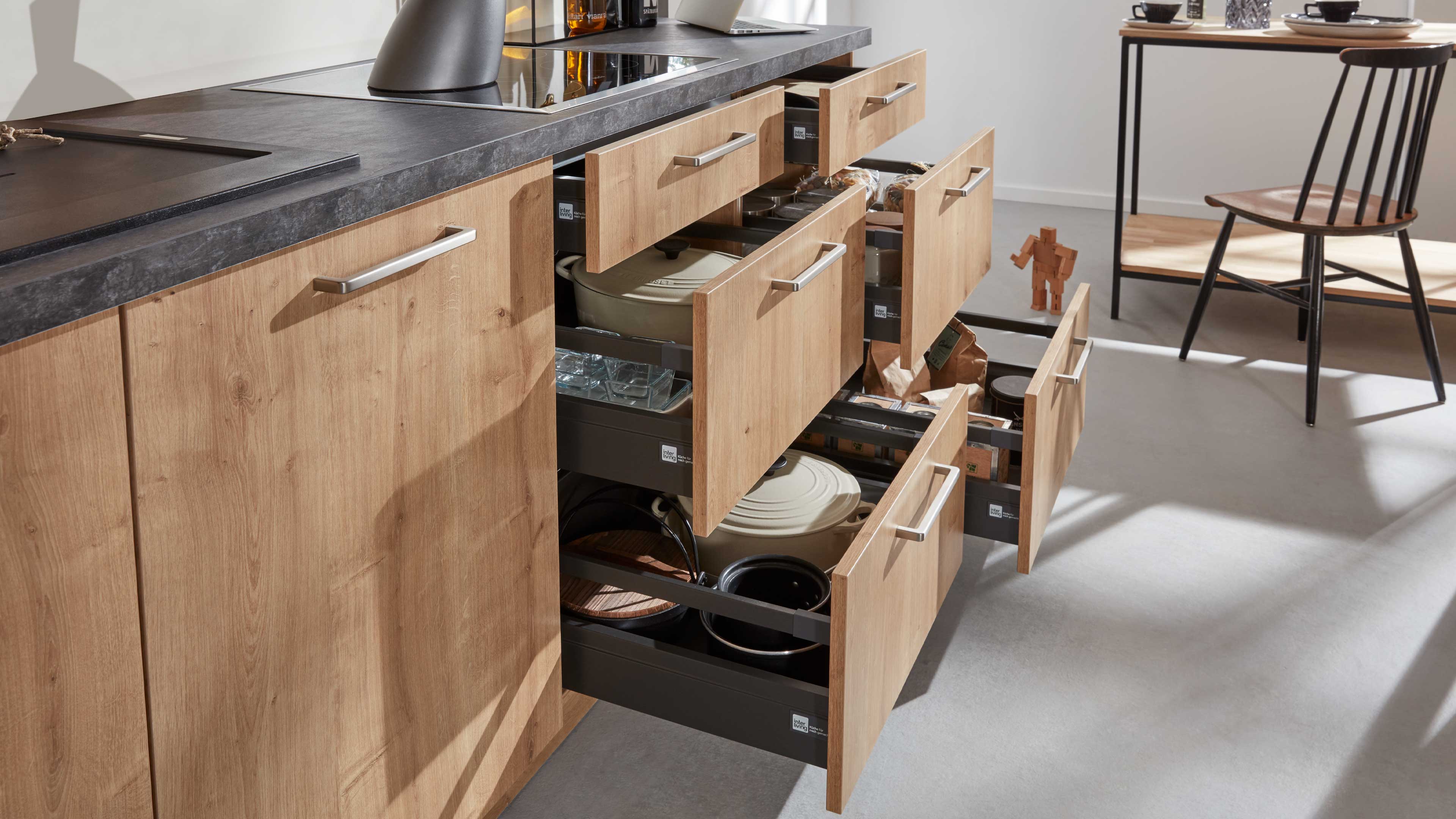 Küchenunterschrank mit hellen Holzfronten und Metallgriffen, dunkler Arbeitsplatte und mehreren offenen Schubladen mit Kochgeschirr.
