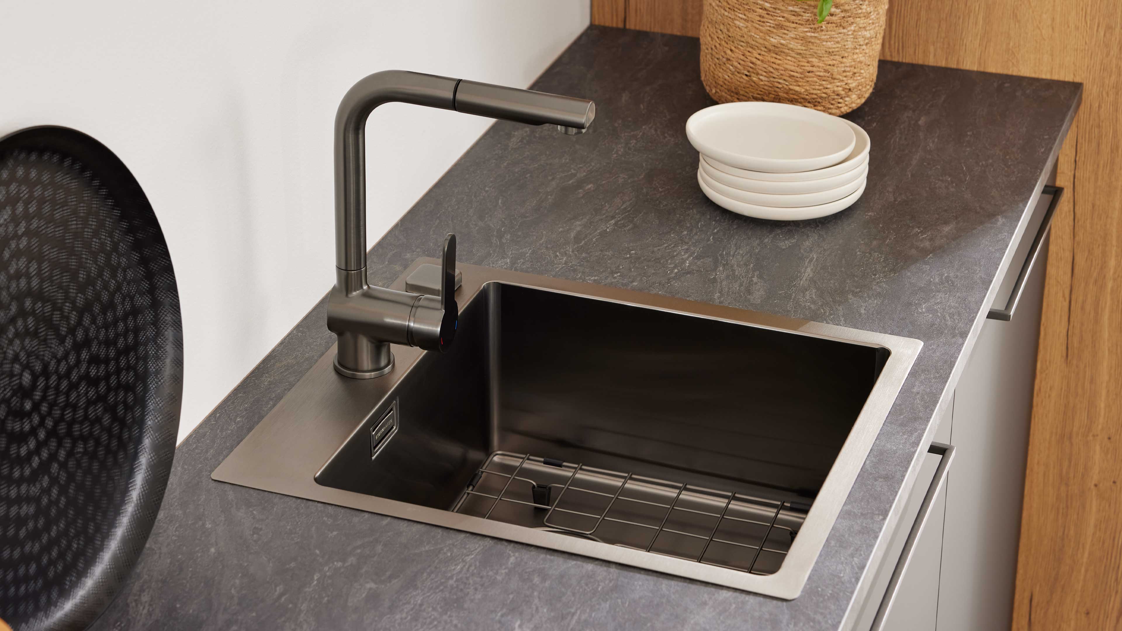Rechteckiges Edelstahlspülbecken mit einarmigem, dunkelgrauem Wasserhahn auf grauer Arbeitsplatte, daneben weiße Schalen und ein Korb.