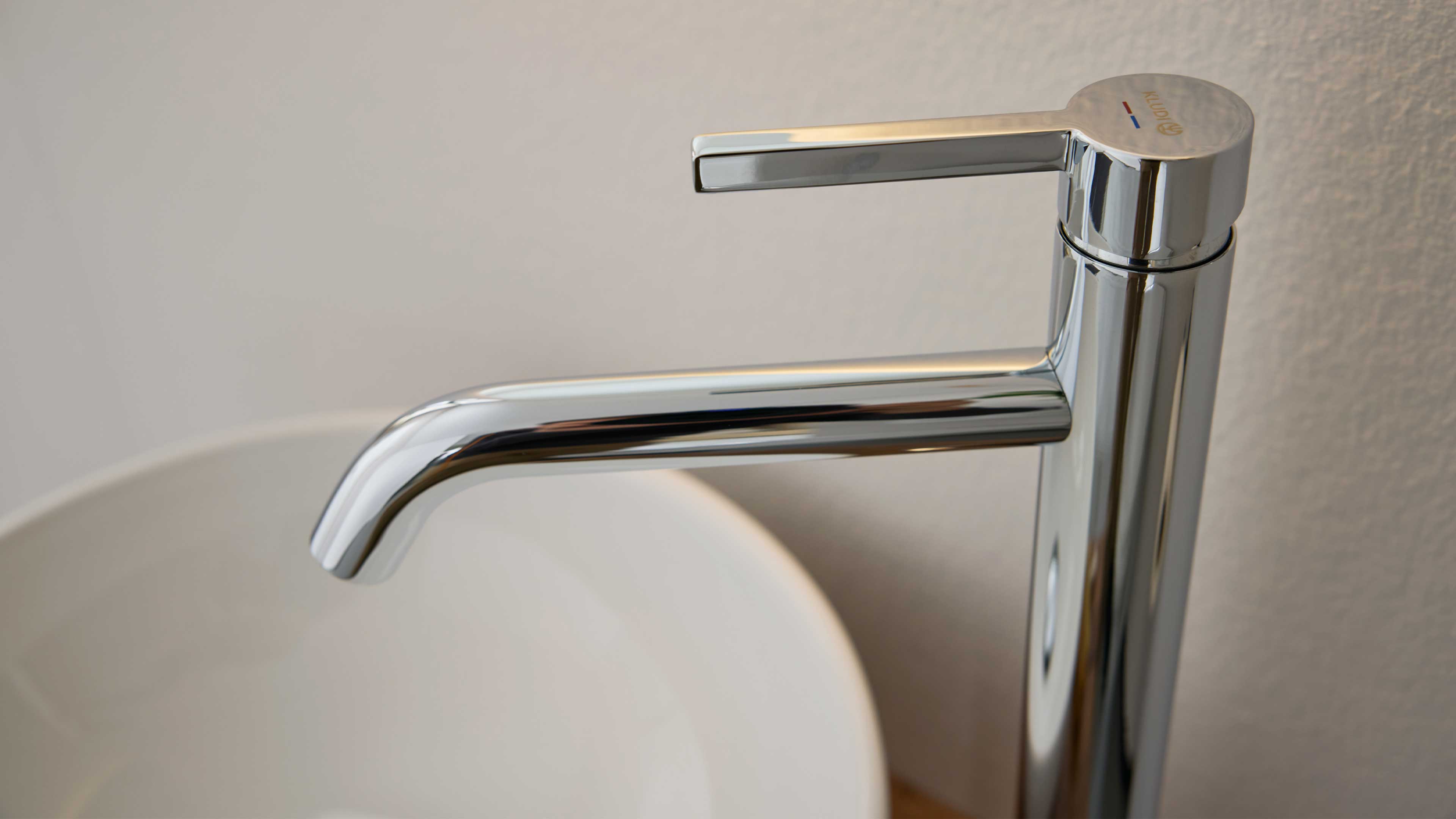 Chromarmatur mit oben liegendem Einhebel, langer gebogener Auslauf über einem weißen Aufsatzwaschbecken, hellgraue Wand im Hintergrund.