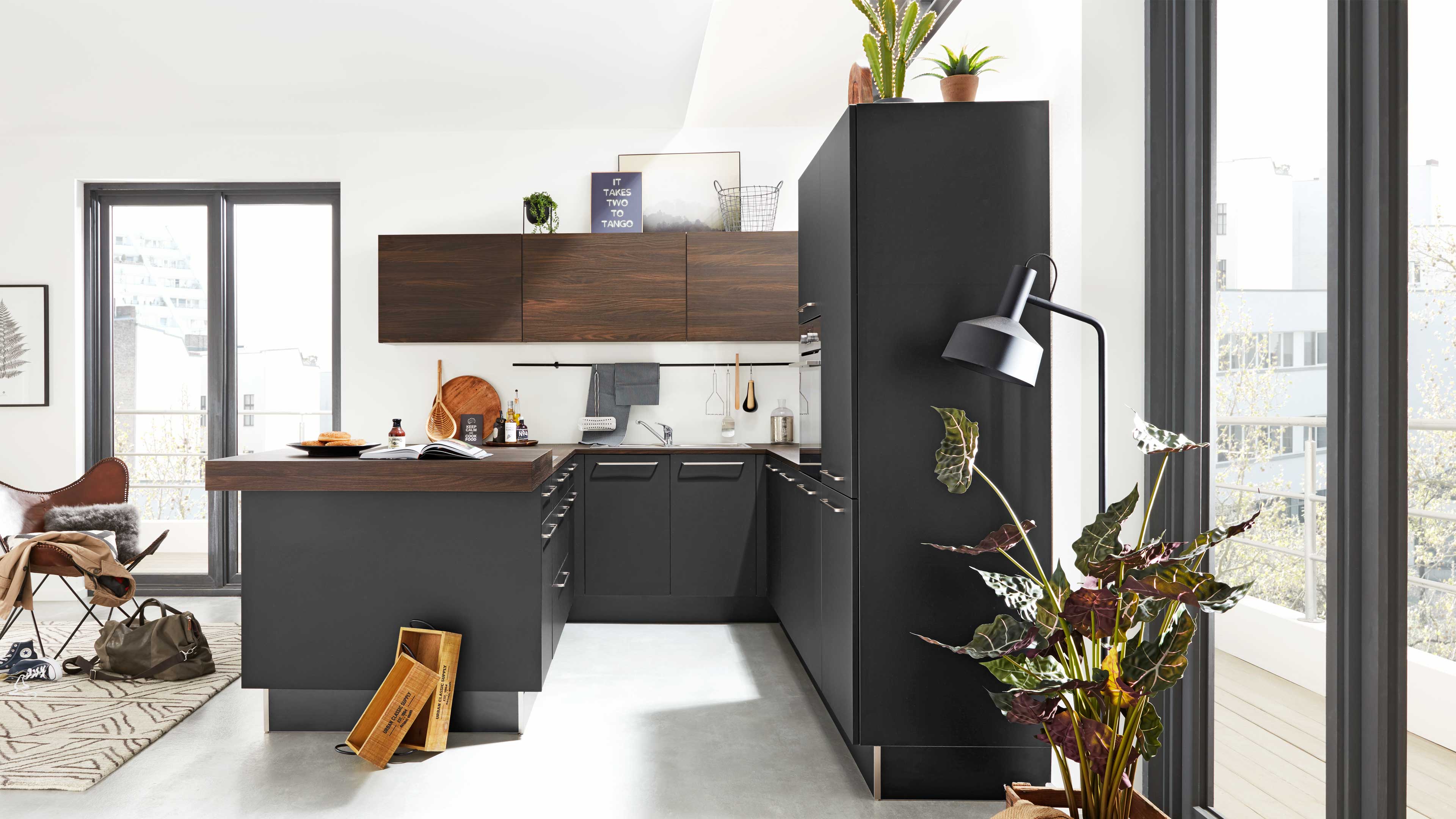 Dunkle Küchenzeile mit Insel und Holzarbeitsplatte, dazu hohe dunkle Schränke, hängende Holzwandschränke und Zimmerpflanze am Fenster.