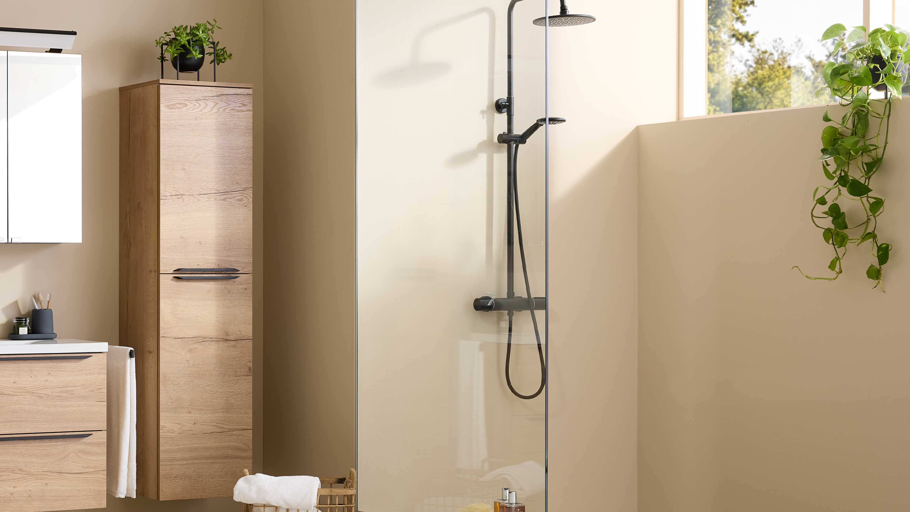 Wandhängender schmaler Hochschrank in heller Holzoptik mit zwei Griffen links, daneben Glas-Duschwand, Körbchen und hängende Grünpflanze.