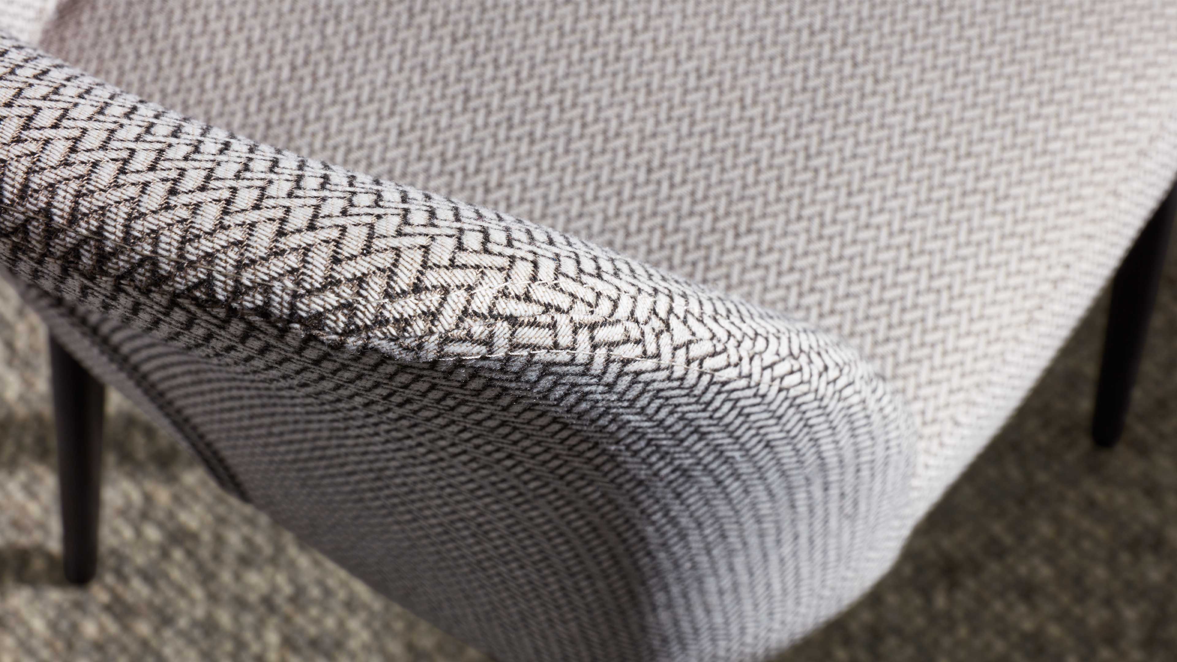 Gepolsterte Armlehne und Sitzfläche eines Stuhls mit dicht gewebtem hellgrau-beigem Fischgrätmuster und sichtbaren schwarzen Metallbeinen.