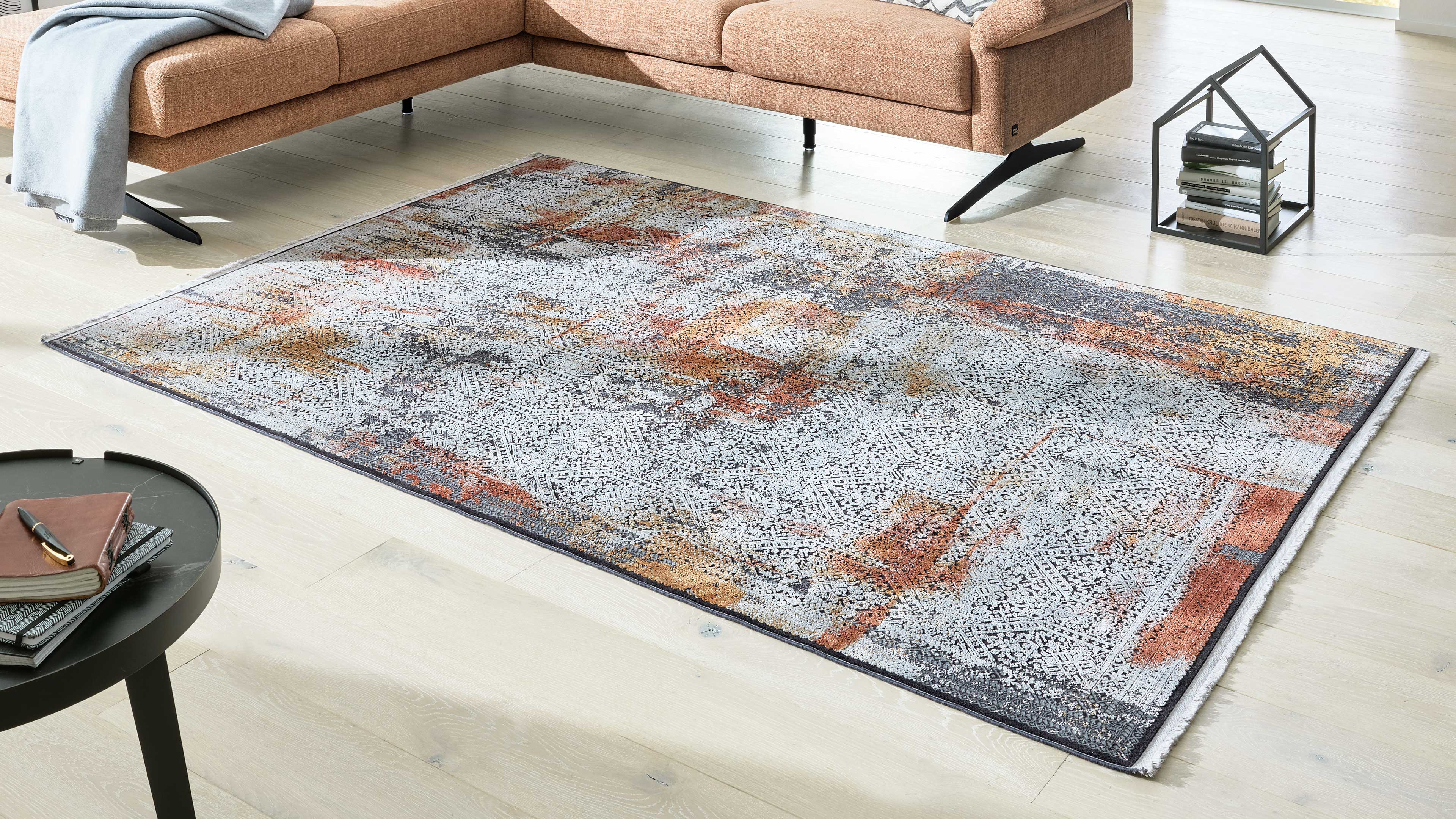 Großflächiger, gemusterter Teppich in Weiß-, Orange- und Grautönen auf hellem Parkett, daneben Ecksofa, Beistelltisch und kleines Bücherregal.