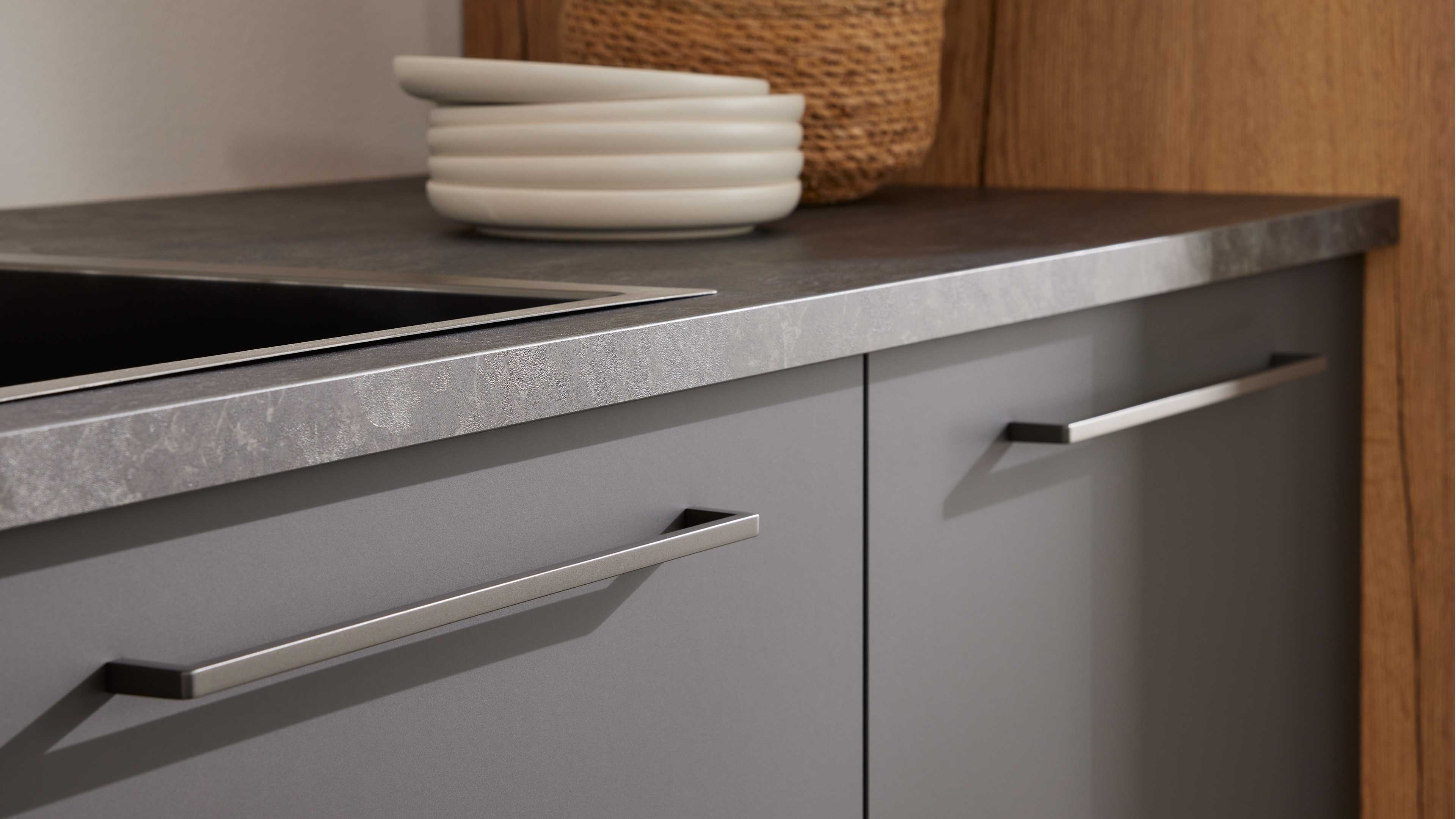 Graue Küchenschrankfronten mit langen Metallgriffen unter einer grauen, steinoptik Arbeitsplatte, oben weiße Tellerstapel.
